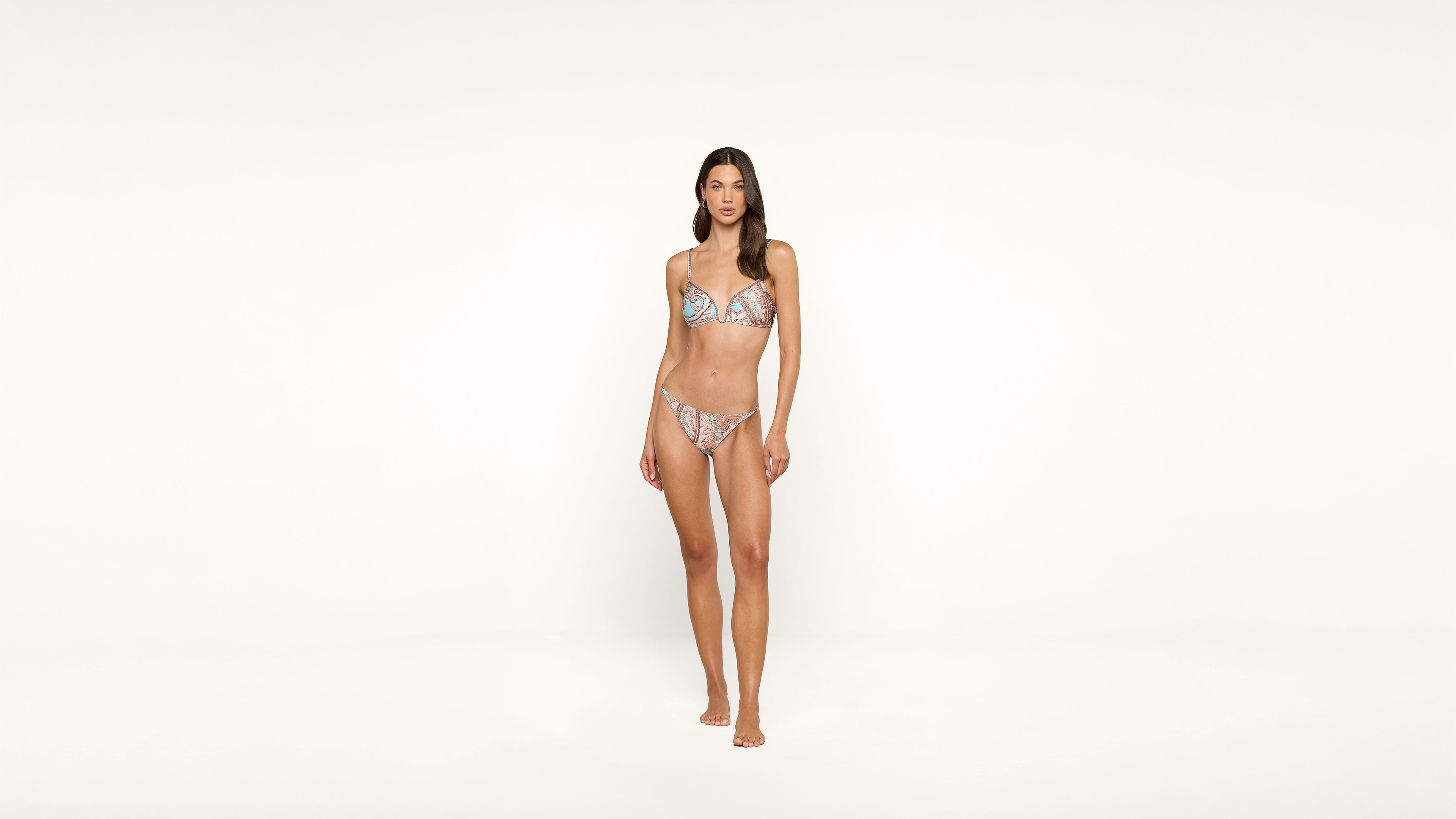 A woman stands barefoot against a plain white background, wearing a patterned bikini.