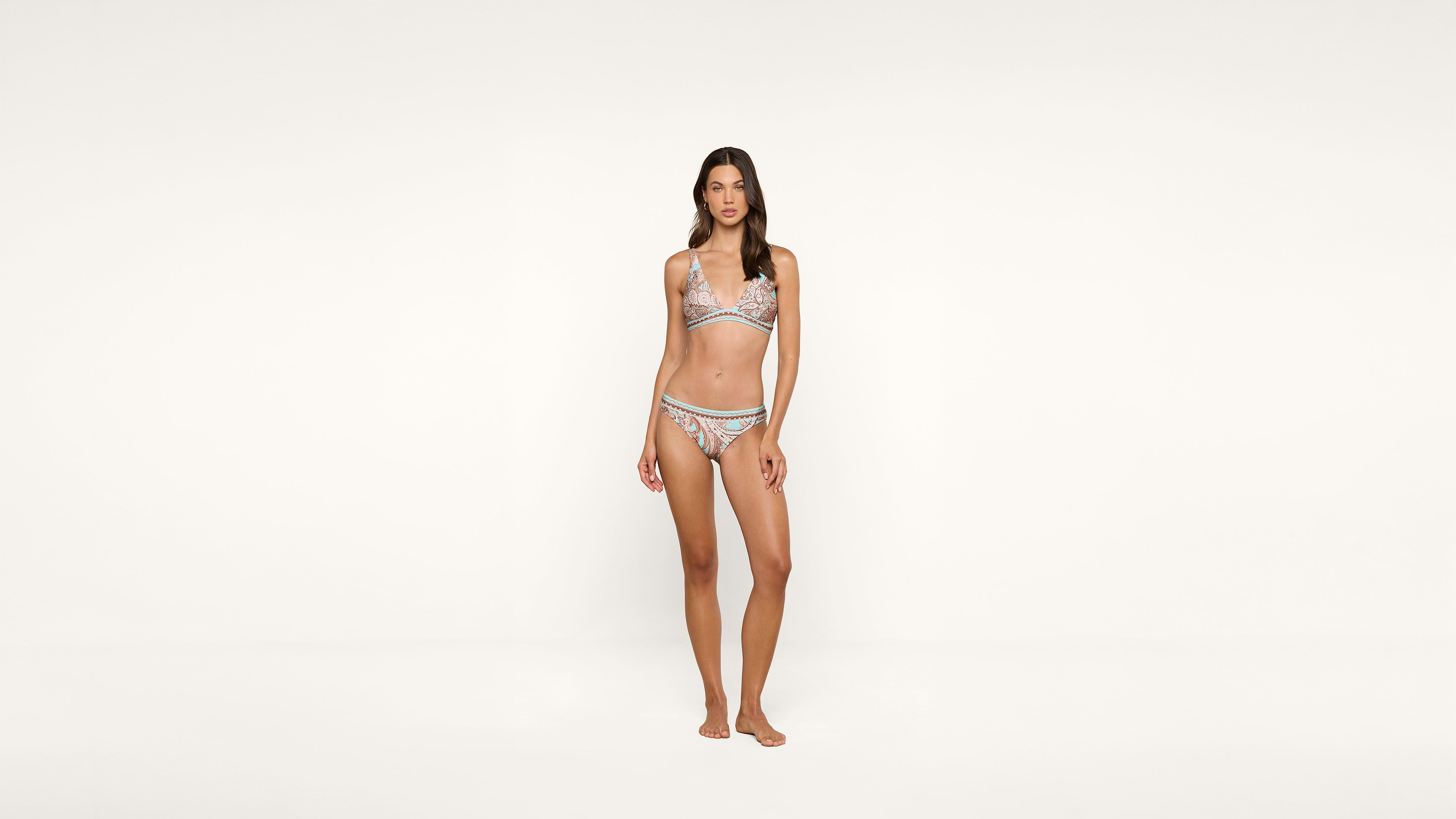 A woman stands barefoot against a plain white background, wearing a patterned bikini. She faces forward with her arms relaxed at her sides.