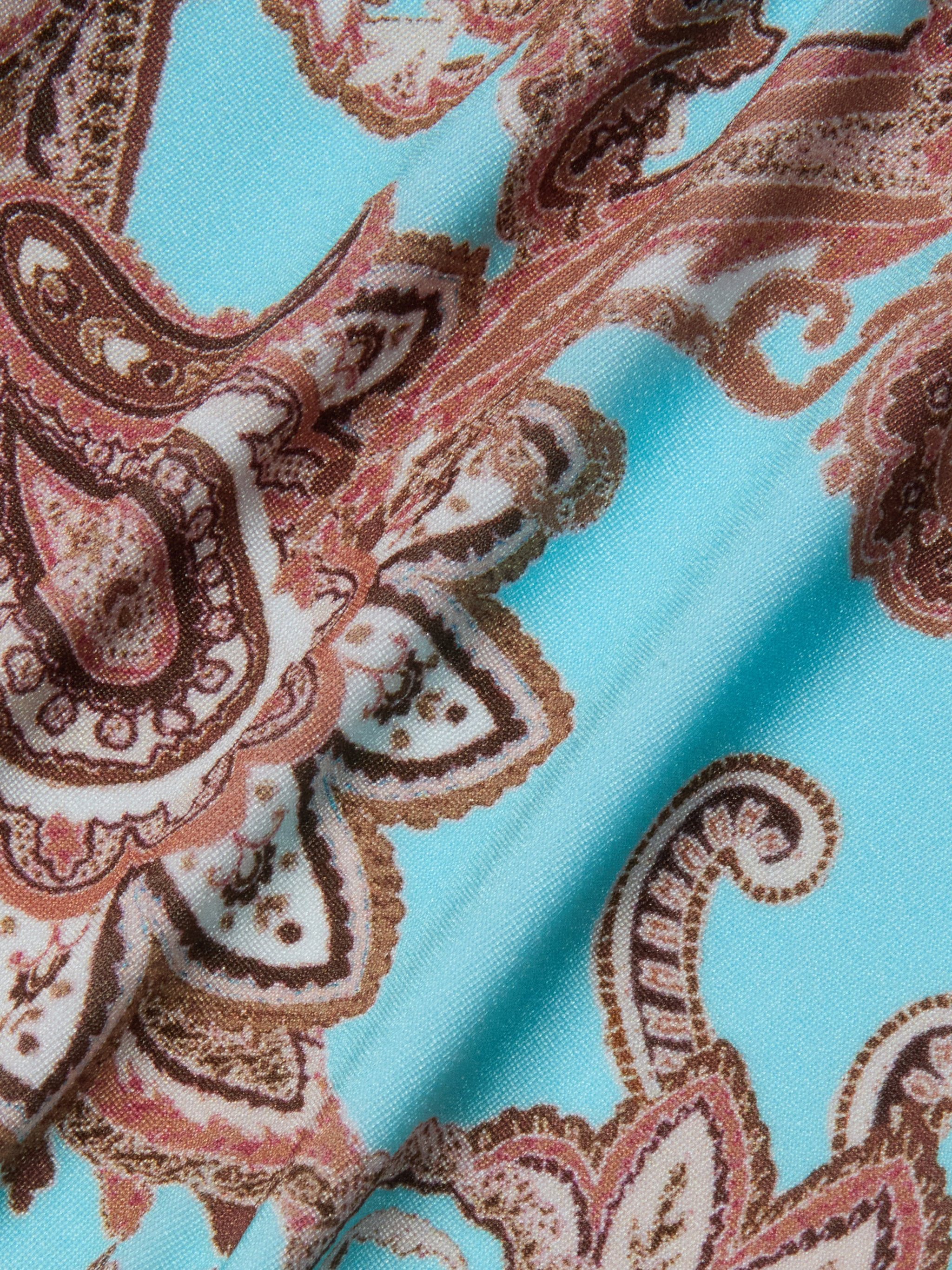 A close-up of the Violet String Bikini Bottom showcases light blue fabric adorned with a paisley pattern in brown, cream, and white. The swirling shapes and intricate details are reminiscent of a classic string bikini bottom, with the fabric appearing slightly folded.