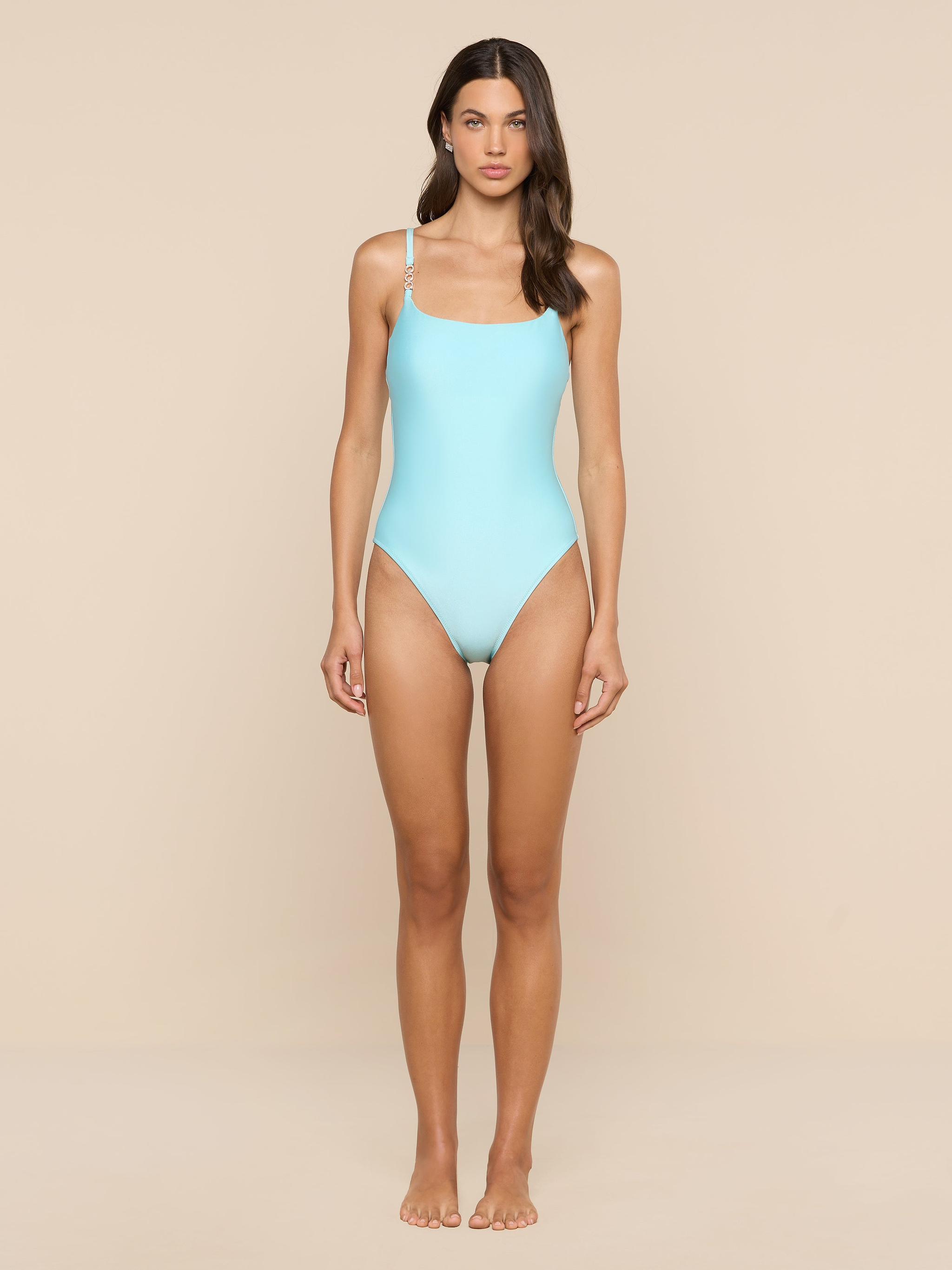 A woman stands barefoot against a beige background, wearing the Remi One-Piece Swimsuit in ice blue with thin straps. She faces forward with a neutral expression and her hair loose over one shoulder.