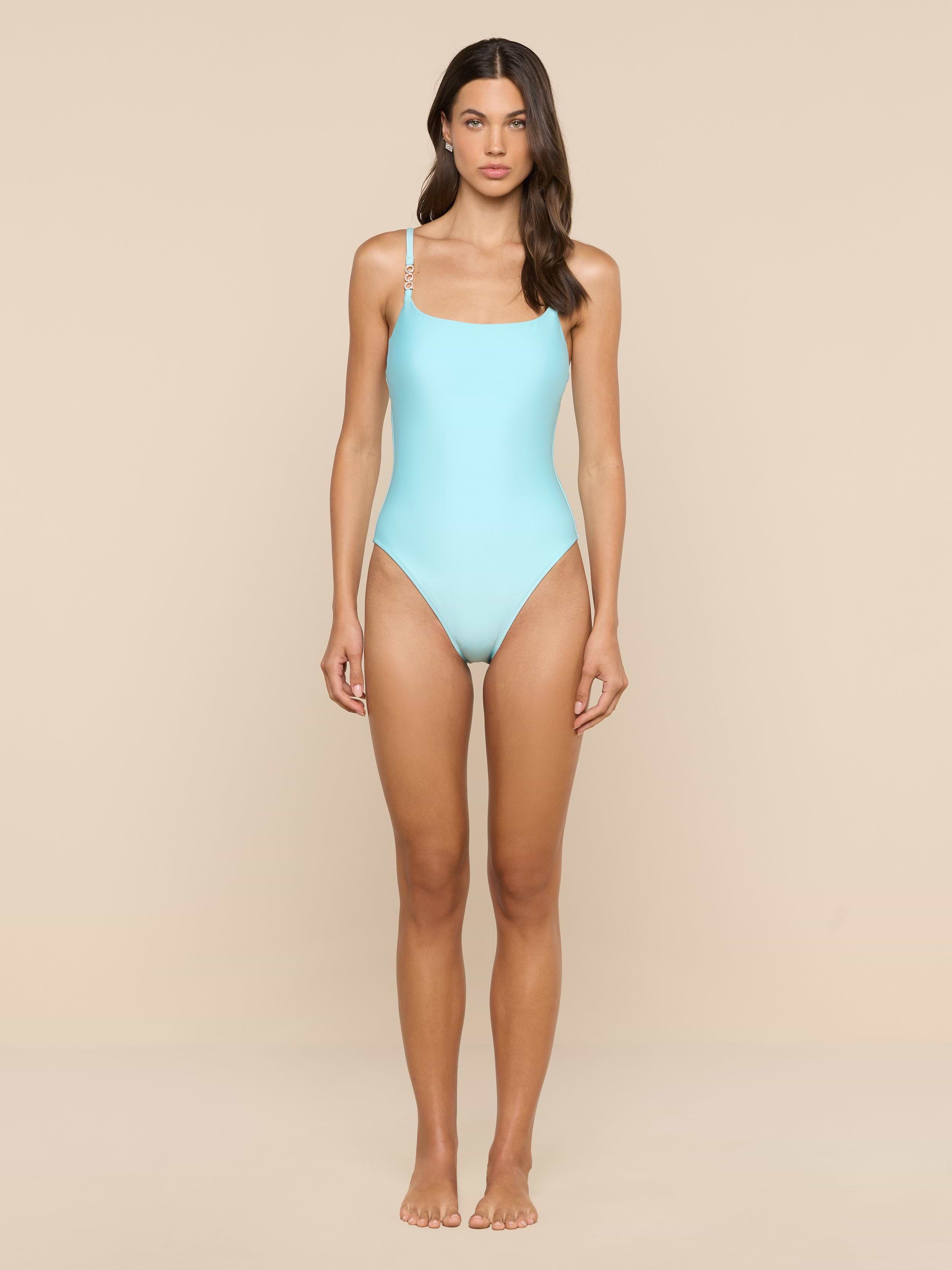 A woman stands barefoot against a beige background, wearing the Remi One-Piece Swimsuit in ice blue with thin straps. She faces forward with a neutral expression and her hair loose over one shoulder.