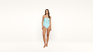 A woman stands barefoot against a plain white background, wearing a light blue strapless one-piece swimsuit with a front lace-up detail.