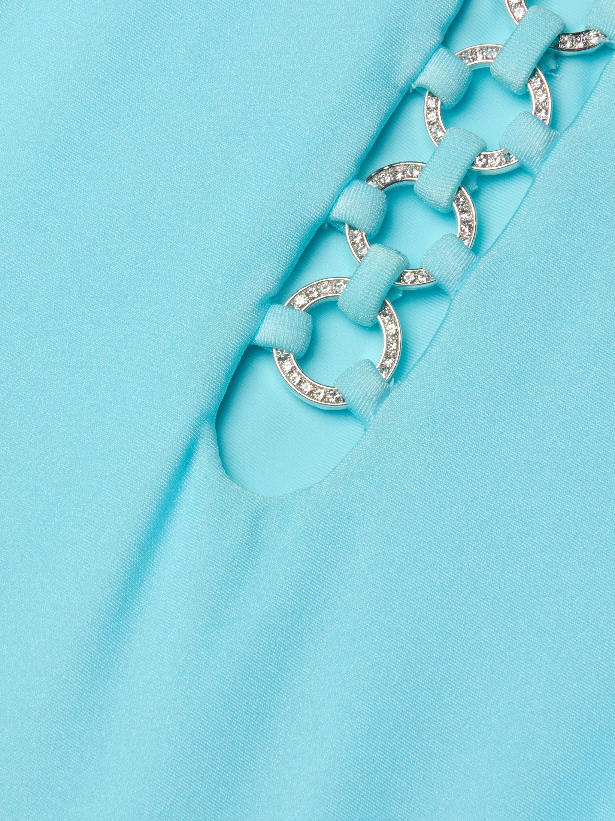 Close-up of the Indore One-Piece Swimsuit in ice blue, showcasing fabric with a cutout detail. Silver rings, linked by fabric loops, accentuate the design—some adorned with small, sparkling rhinestones.