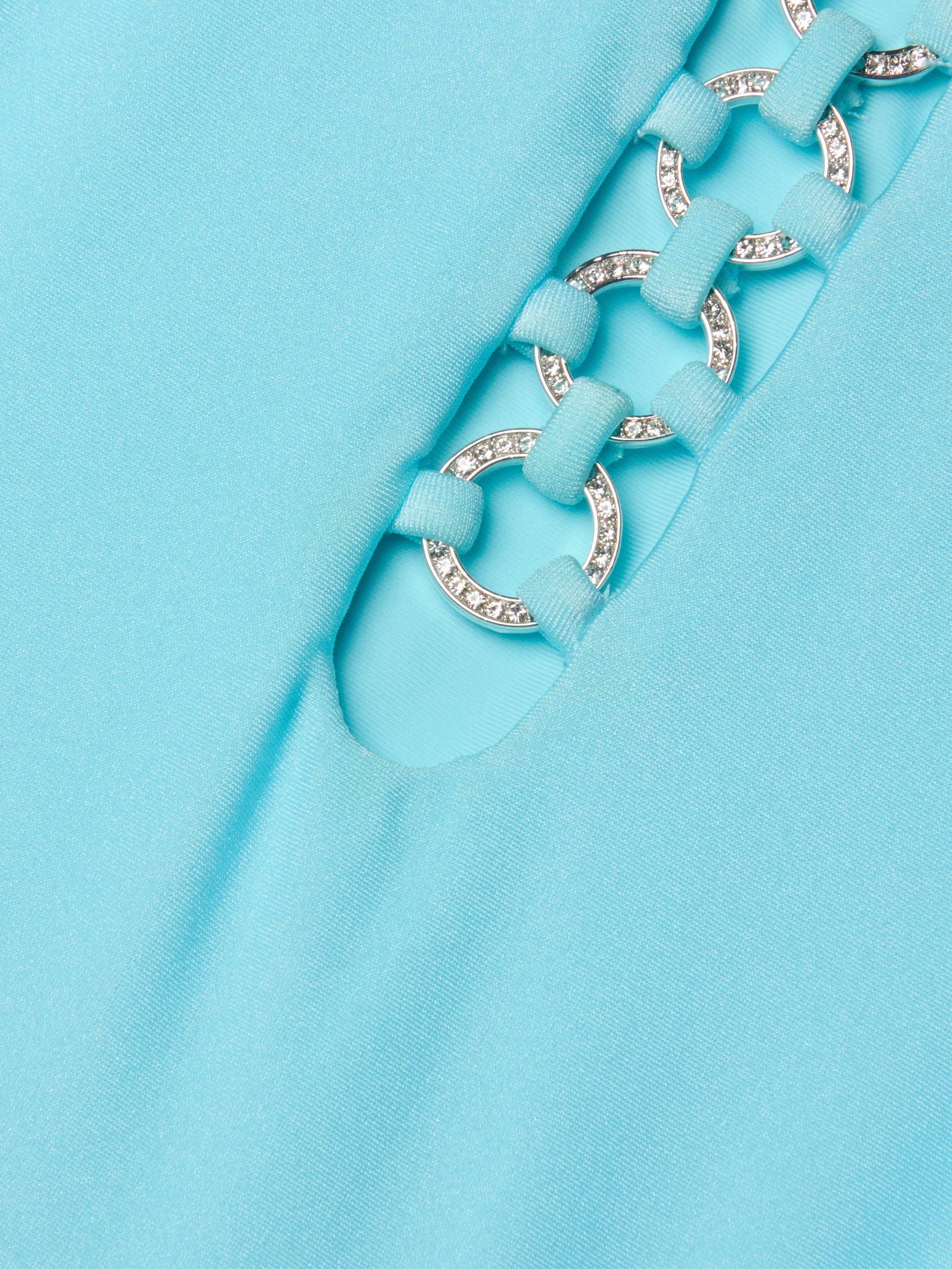 Close-up of the Indore One-Piece Swimsuit in ice blue, showcasing fabric with a cutout detail. Silver rings, linked by fabric loops, accentuate the design—some adorned with small, sparkling rhinestones.