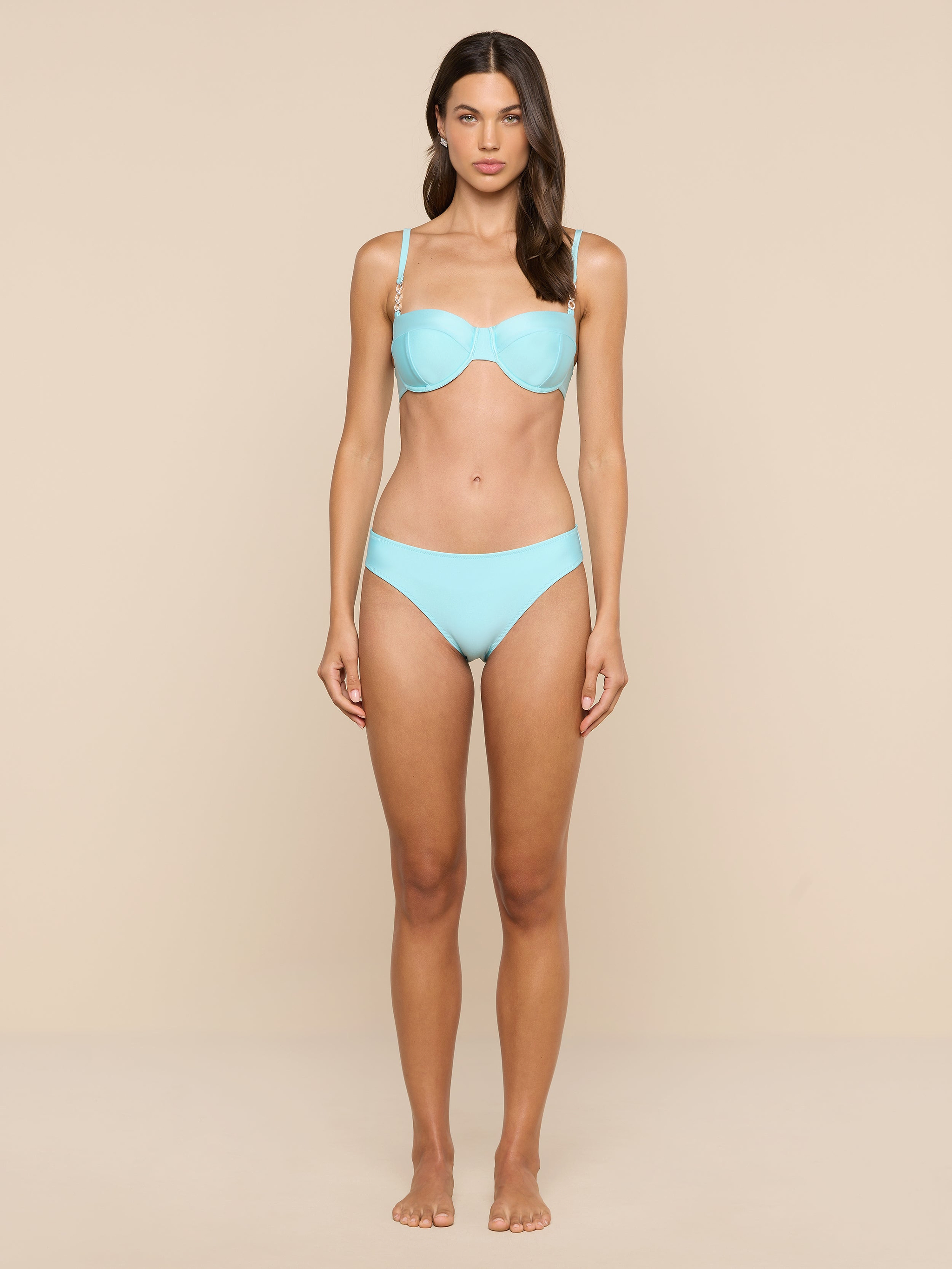 A woman stands barefoot facing forward, wearing an ice blue Alexandria Bikini Top as part of her bikini set, against a plain white background.