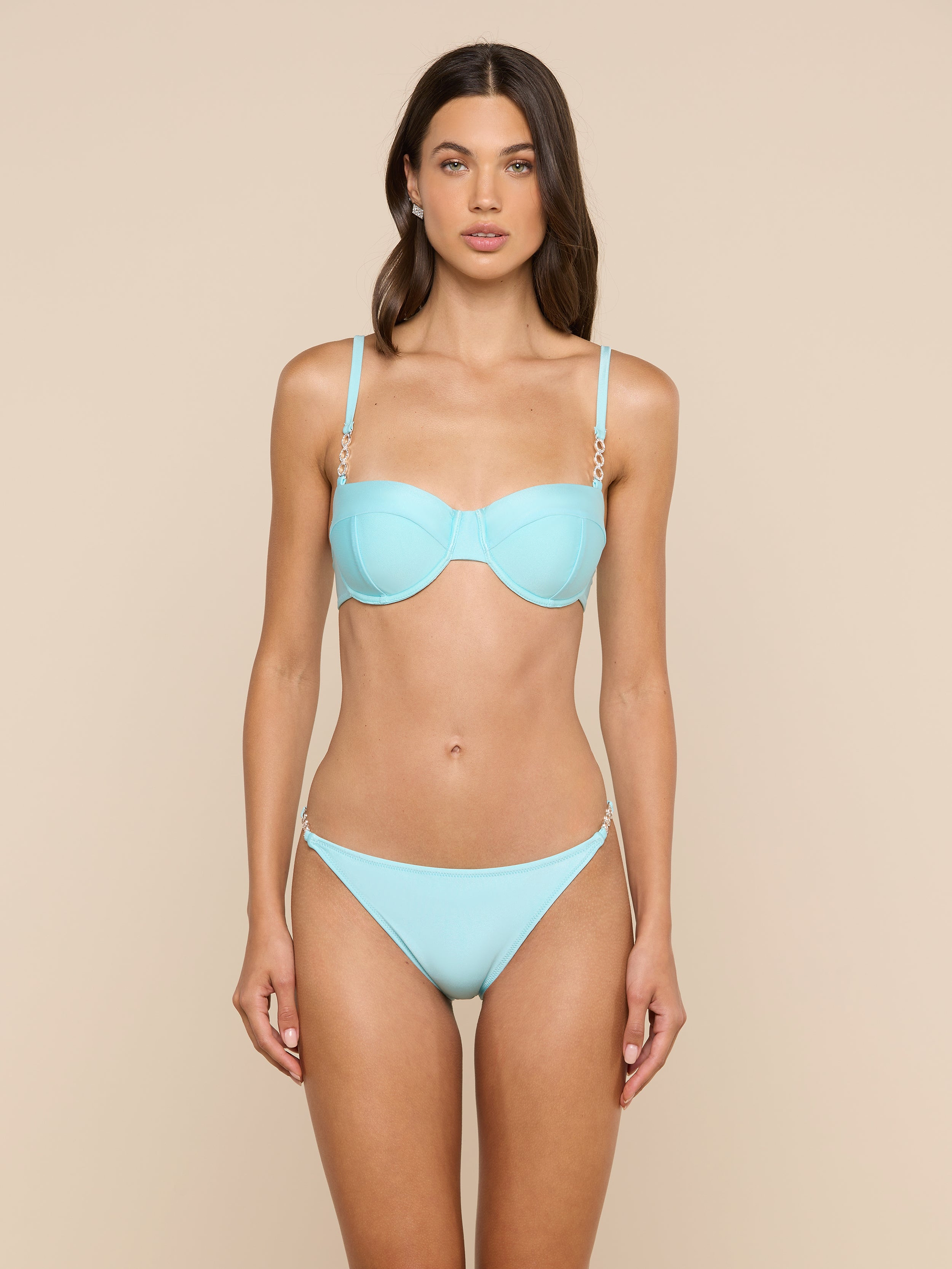 A woman stands facing forward, wearing the Violet String Bikini Bottom in ice blue with gold chain details at the hips, against a plain, light background.