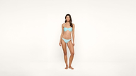 A woman stands barefoot against a plain white background, wearing a light blue bikini and looking directly at the camera with a neutral expression.