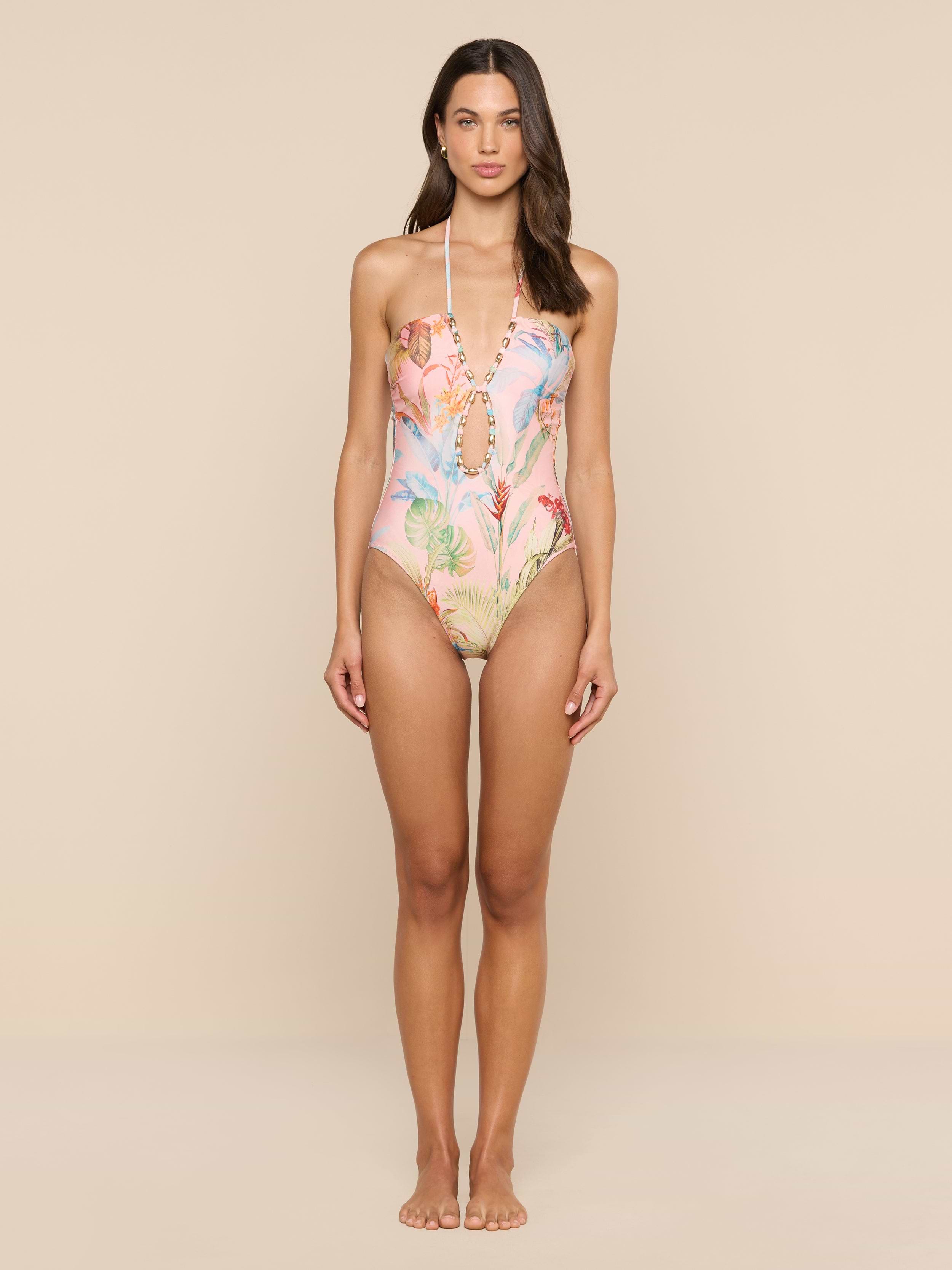 A woman stands against a plain white background, wearing the Opus Halter One-Piece Swimsuit, which features a modern colorful floral and coral reef pattern. She faces forward with her arms relaxed by her sides, showcasing this stylish UPF 50 swimsuit.