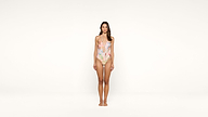 A woman stands barefoot against a plain white background, wearing a pastel floral one-piece swimsuit. She faces forward with arms relaxed at her sides.