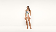 A woman stands barefoot against a plain white background, wearing a colorful one-piece floral swimsuit. She faces forward with a relaxed expression and her arms at her sides.