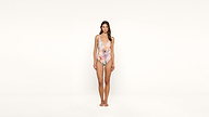 A woman stands barefoot against a plain white background, wearing a floral one-piece swimsuit with thin straps, looking directly at the camera.