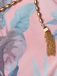 Gold beaded necklace with a chain tassel lying on pink fabric featuring blue and green leafy patterns, reminiscent of the vibrant hues in the Breana Plunge One-Piece Swimsuit.