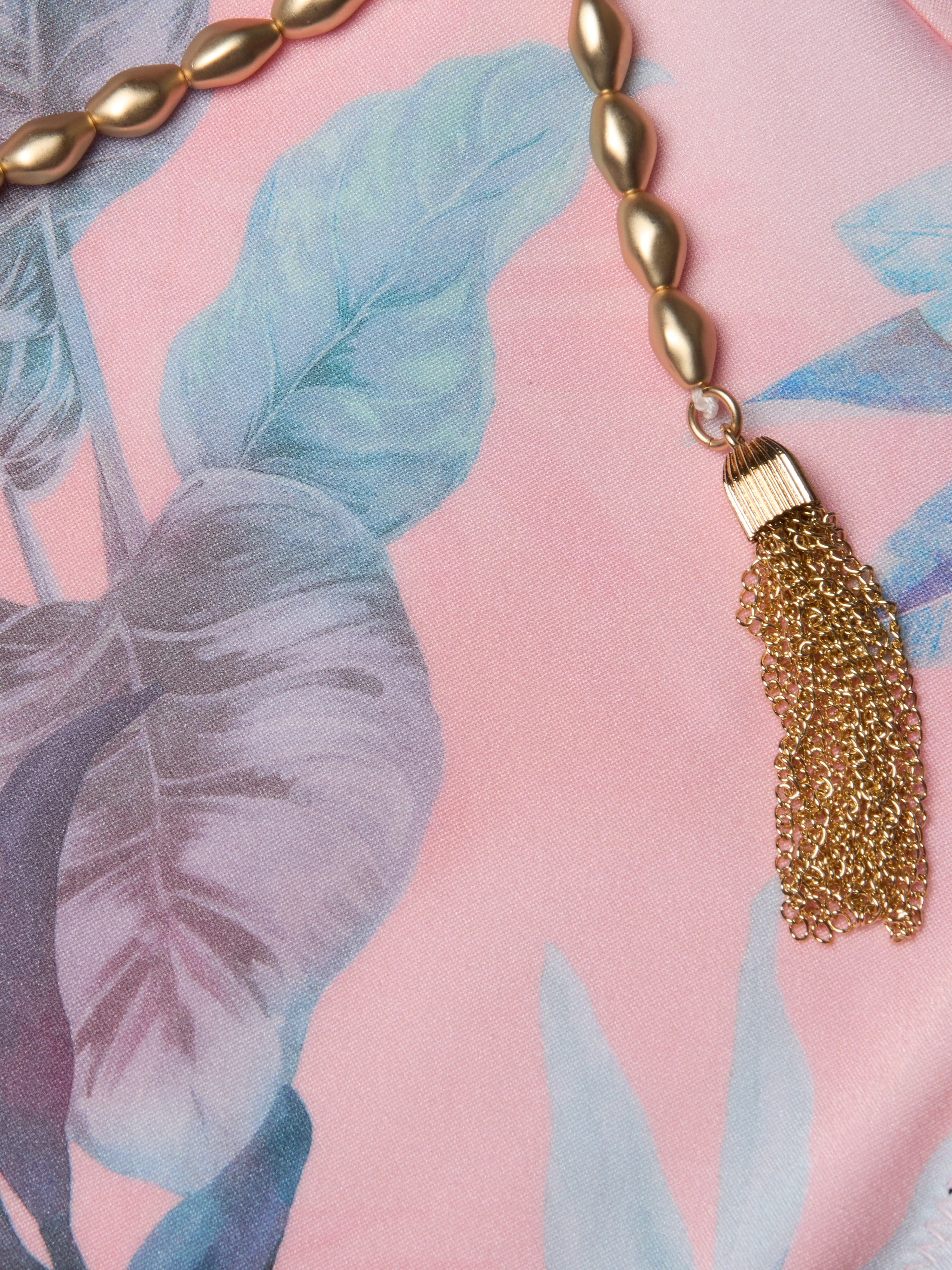Gold beaded necklace with a chain tassel lying on pink fabric featuring blue and green leafy patterns, reminiscent of the vibrant hues in the Breana Plunge One-Piece Swimsuit.