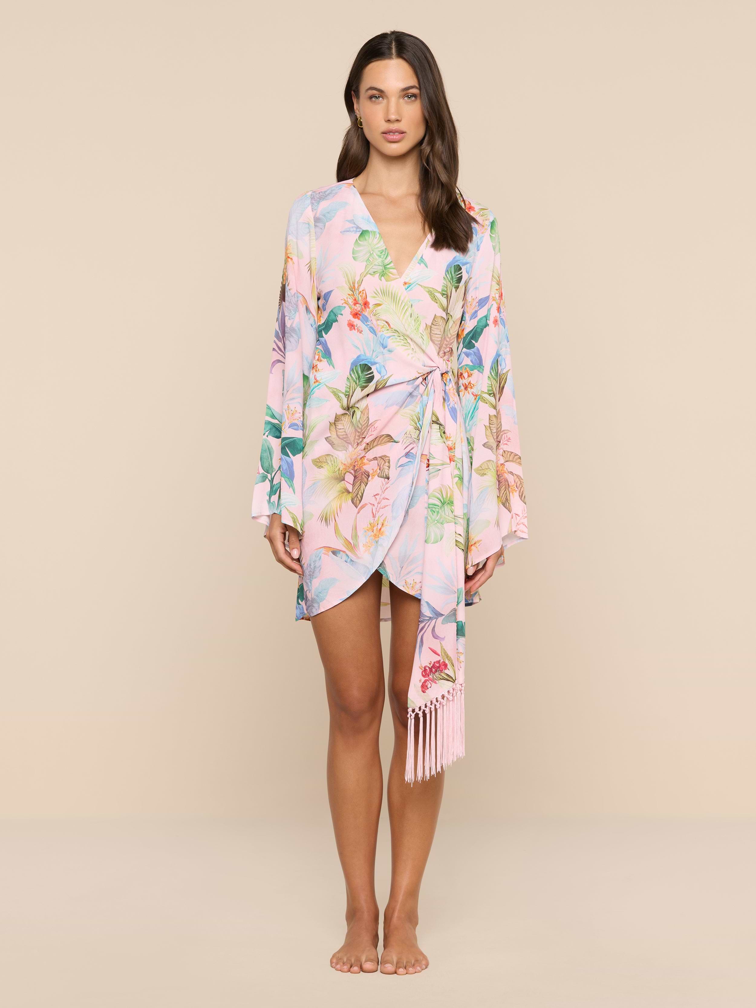 A woman stands facing forward, wearing the Kohima Cover-Up Dress—a pastel floral wrap dress with bell sleeves and a tie at the waist—against a plain white background.