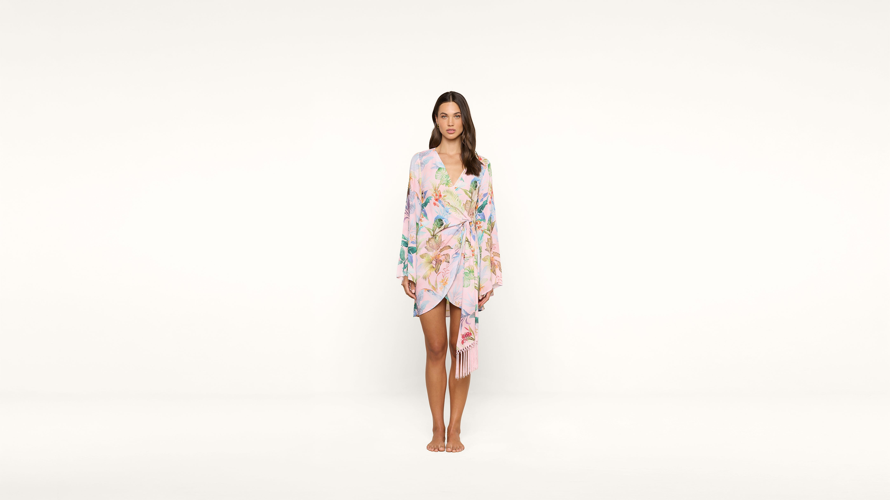 A woman stands barefoot against a plain white background, wearing a light pink floral kimono-style wrap dress with long sleeves and a tied waist.