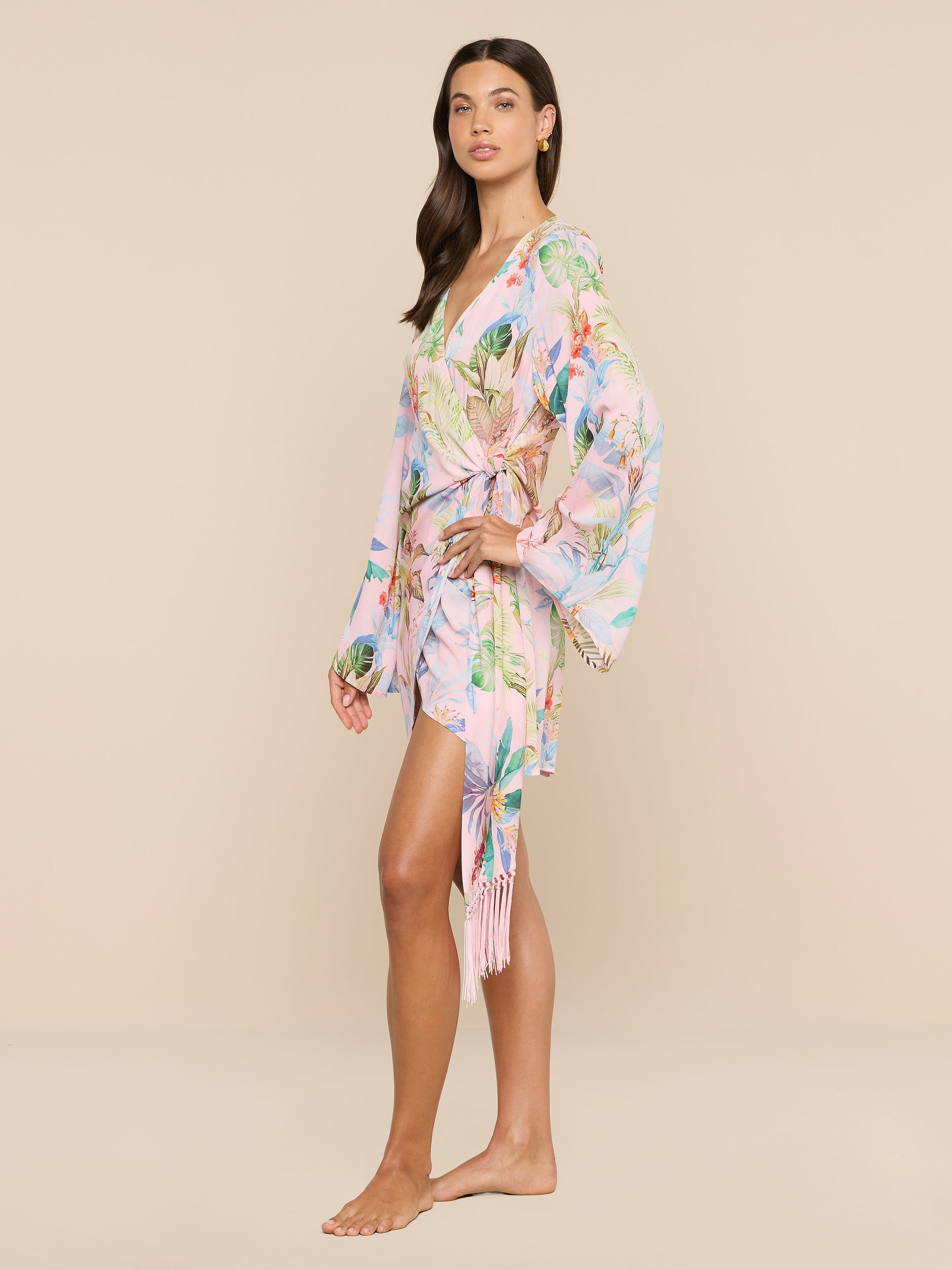 A woman stands barefoot in the Kohima Cover-Up Dress, a light pink floral kimono-style piece featuring long, loose bell sleeves and a wrap tie at the waist, evoking effortless resort style against a plain white background.