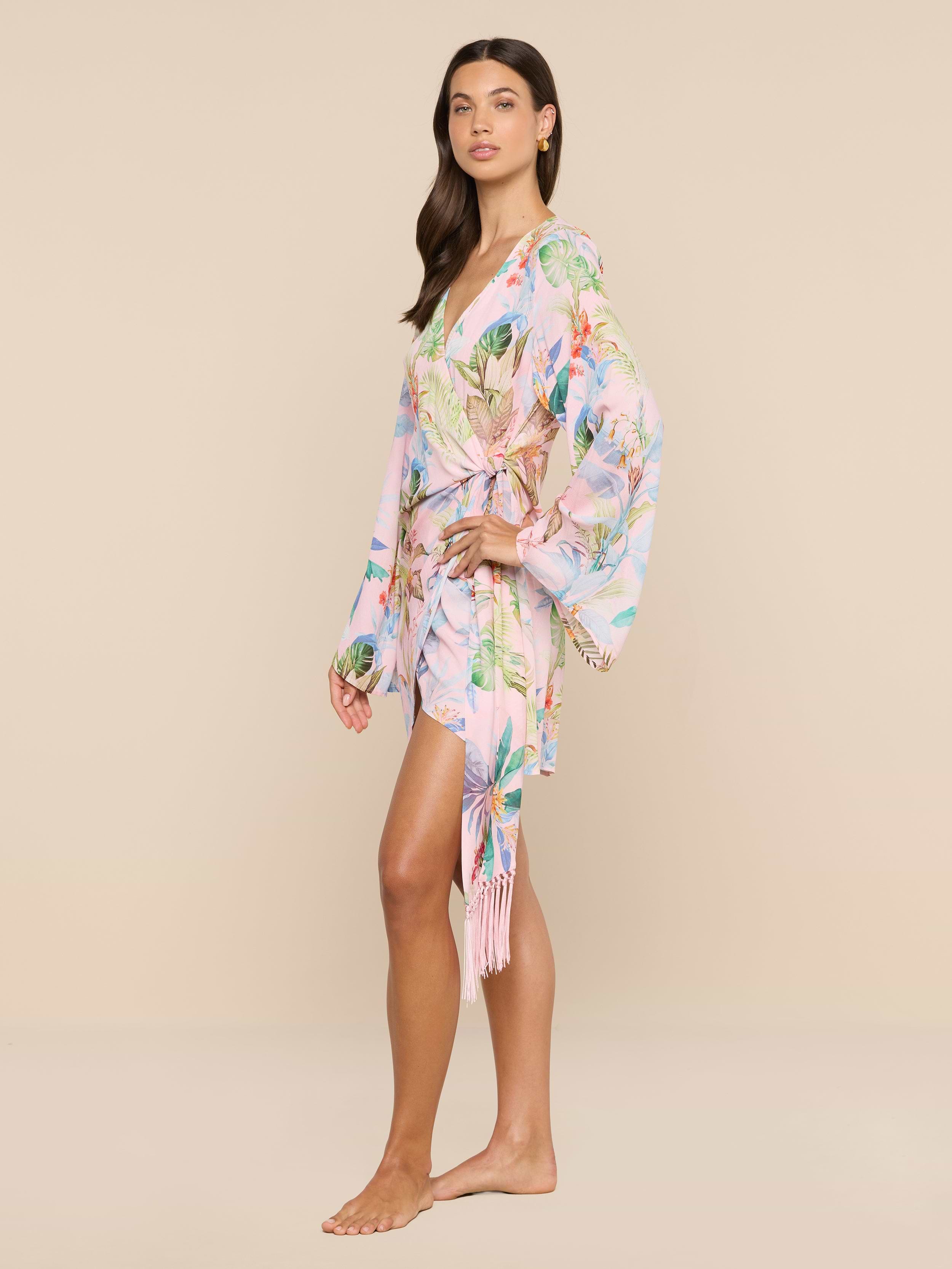 A woman stands barefoot in the Kohima Cover-Up Dress, a light pink floral kimono-style piece featuring long, loose bell sleeves and a wrap tie at the waist, evoking effortless resort style against a plain white background.