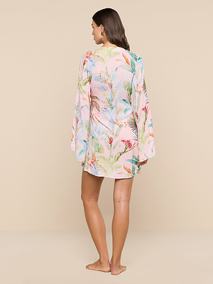 A woman stands barefoot on a beige floor, facing away, wearing the Kohima Cover-Up Dress—a loose resort-style robe in pastel pink with colorful tropical floral and animal prints. Her long brown hair is down against a plain beige background.