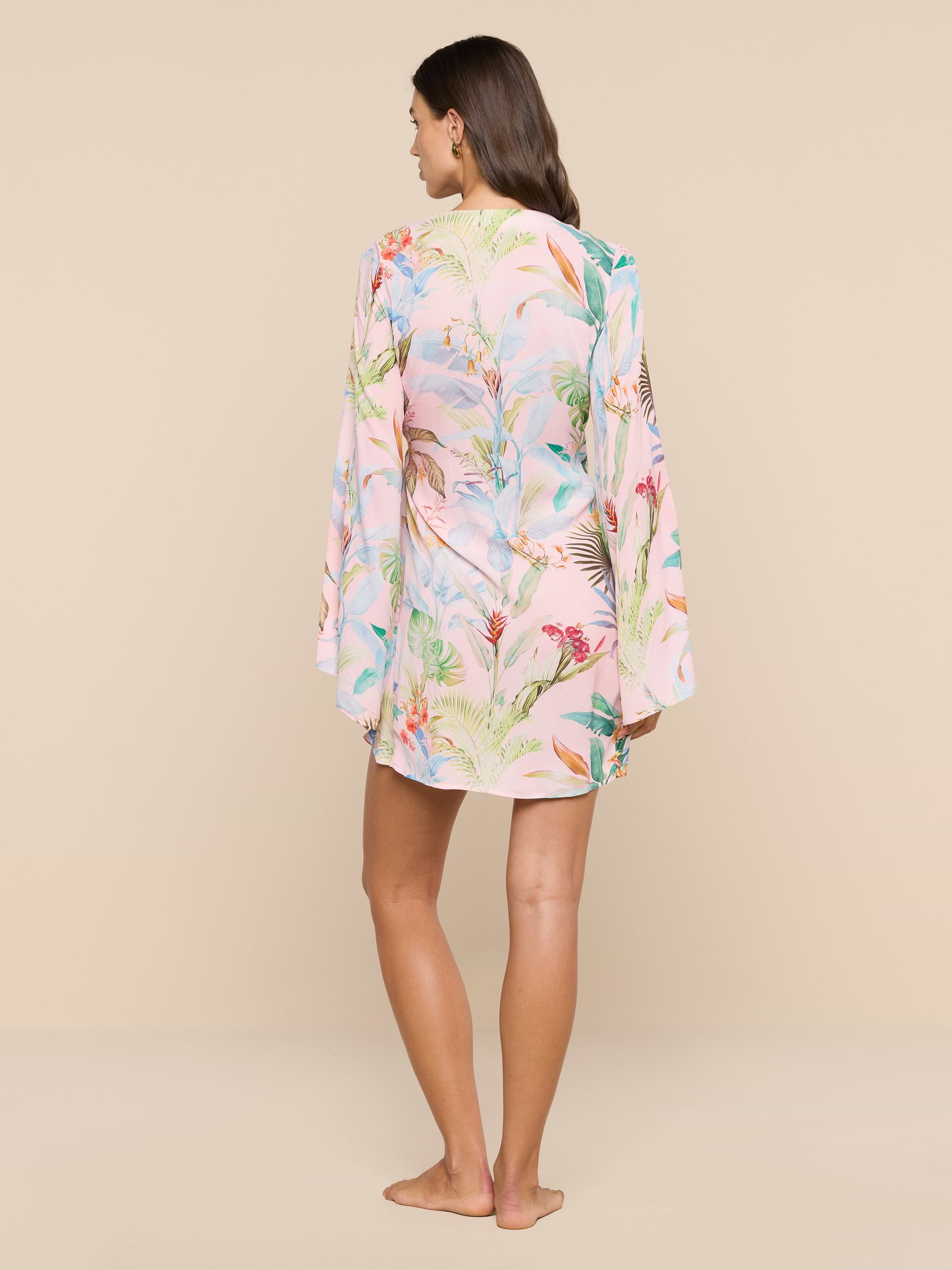 A woman stands barefoot facing away, wearing the Kohima Cover-Up Dress—a light, floral-patterned long-sleeve dress with subtle bell sleeves—set against a plain white background.