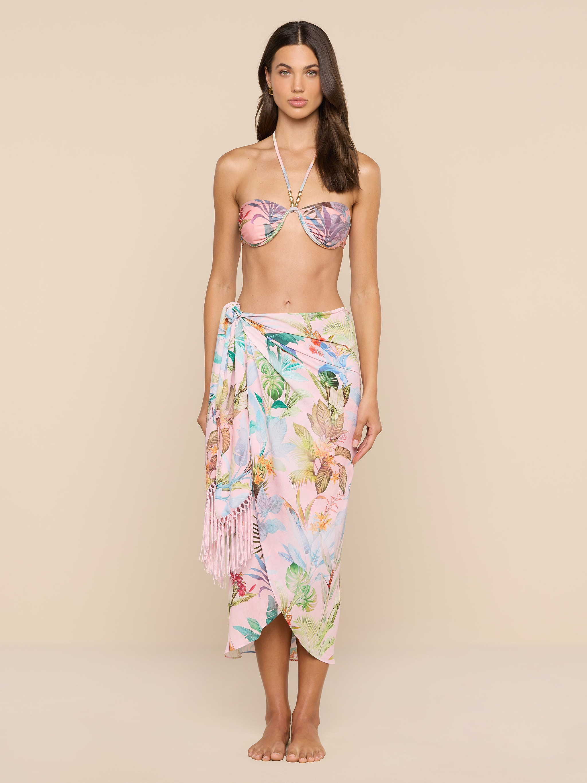 A woman stands barefoot against a beige background, wearing a pink floral bikini top and the Marena Sarong with fringe detail tied at her waist, looking directly at the camera.