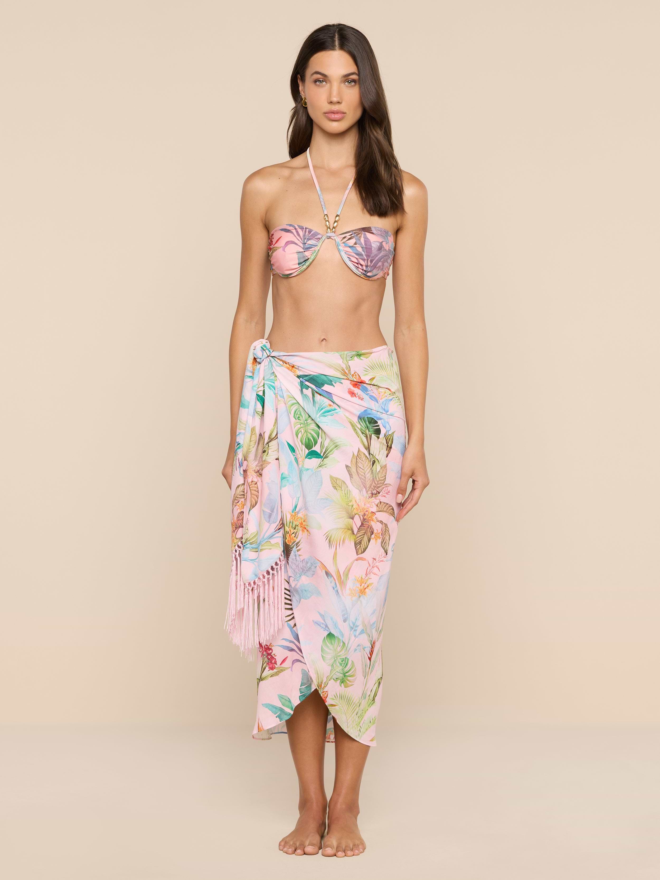 A woman stands barefoot against a beige background, wearing a pink floral bikini top and the Marena Sarong with fringe detail tied at her waist, looking directly at the camera.