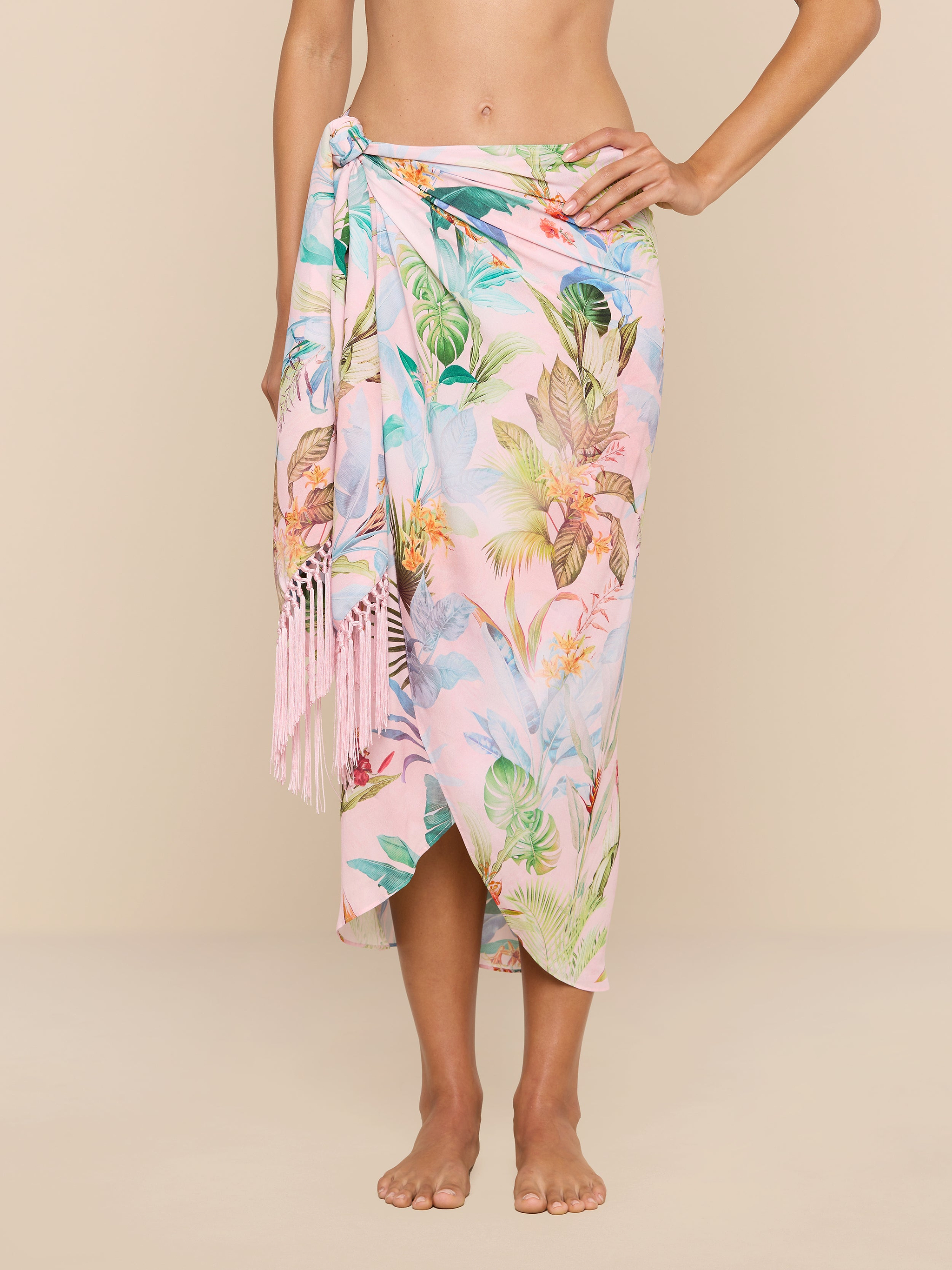 Someone wearing the Marena Sarong in pastel pink with tropical leaf and floral prints, tied at one side with fringe tassels, covering from waist to mid-calf. The background is light beige.