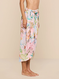 A person stands barefoot against a plain beige background, wearing the Marena Sarong in pastel pink with floral patterns and fringe details, tied at the waist. Only the upper body and feet are visible.
