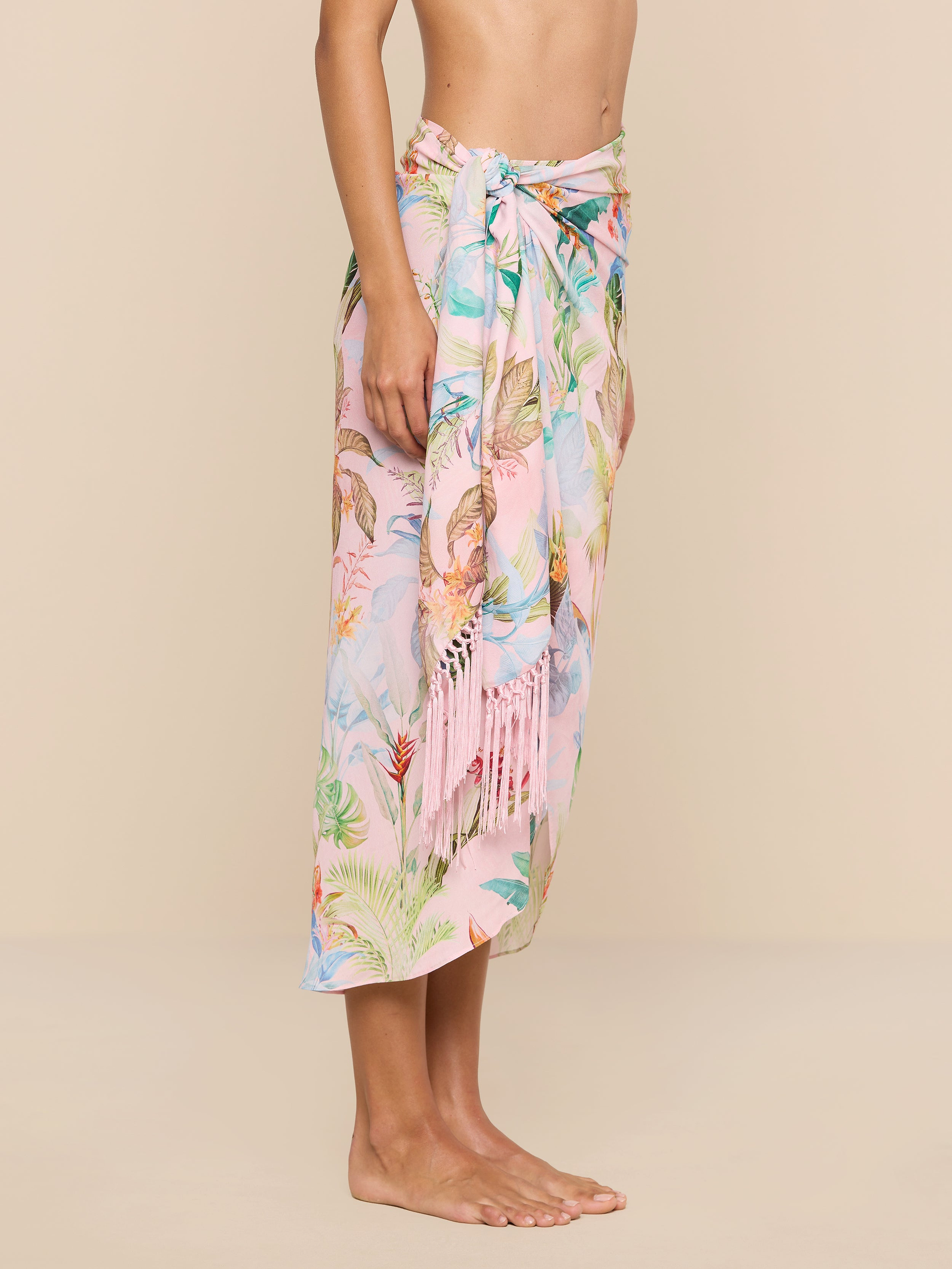 A person stands barefoot against a plain beige background, wearing the Marena Sarong in pastel pink with floral patterns and fringe details, tied at the waist. Only the upper body and feet are visible.