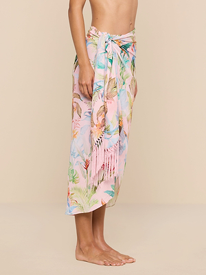 A person stands barefoot against a plain beige background, wearing the Marena Sarong in pastel pink with floral patterns and fringe details, tied at the waist. Only the upper body and feet are visible.