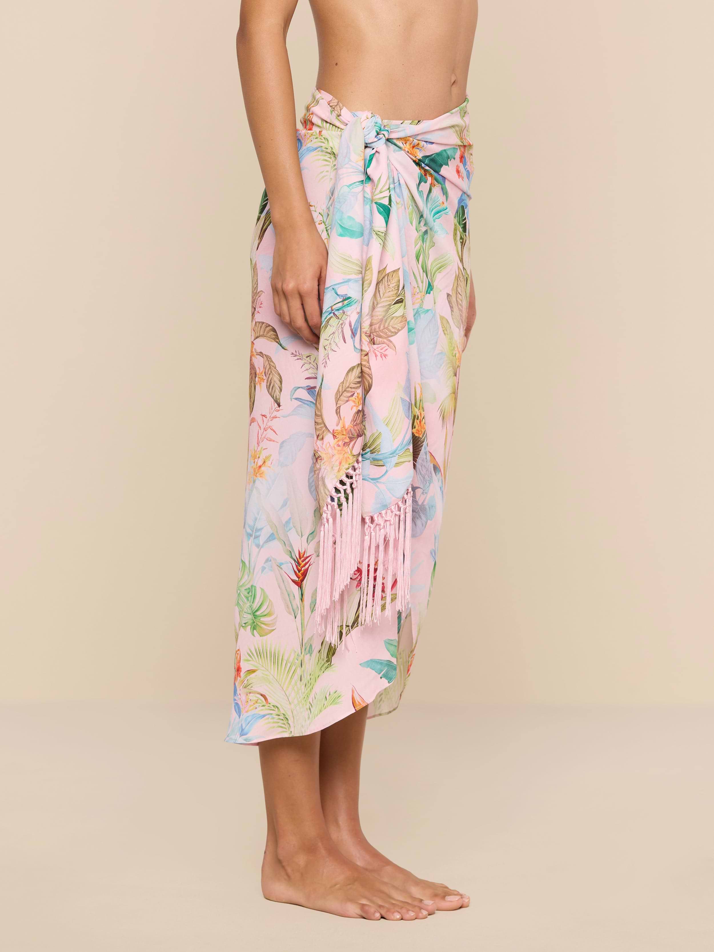 A person stands barefoot against a plain beige background, wearing the Marena Sarong in pastel pink with floral patterns and fringe details, tied at the waist. Only the upper body and feet are visible.