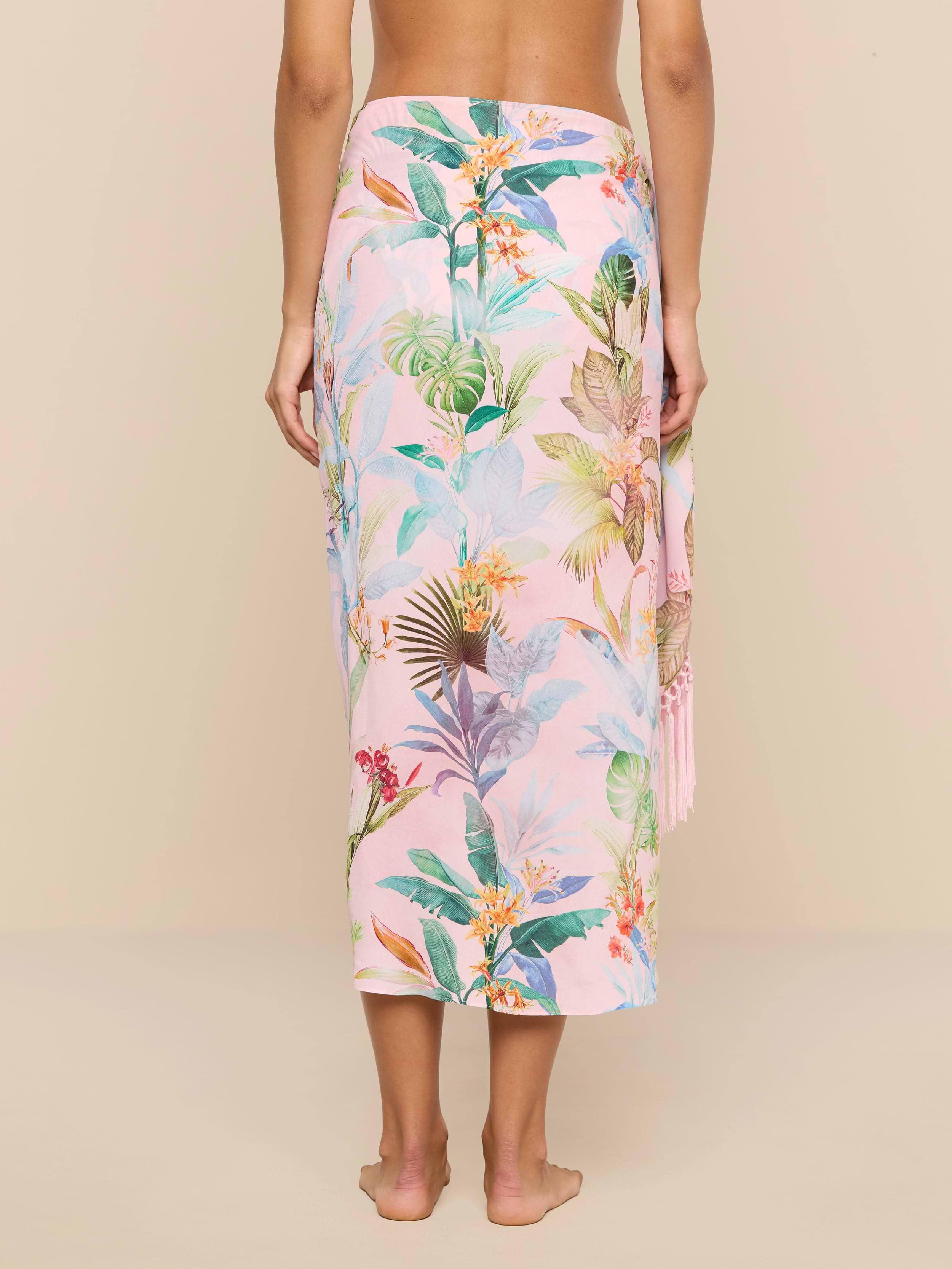 A person stands barefoot facing away, wearing the Marena Sarong in pink with a colorful tropical floral and leaf pattern, set against a plain beige background.
