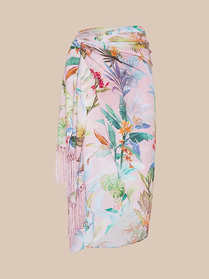 The Marena Sarong is a pastel pink vacation wrap featuring a colorful tropical floral and leaf print, tied at the waist and finished with fringe tassels along one edge, shown against a beige background.