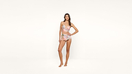 A woman stands barefoot against a plain white background, wearing a colorful floral two-piece outfit with a crop top and shorts, looking confidently at the camera.