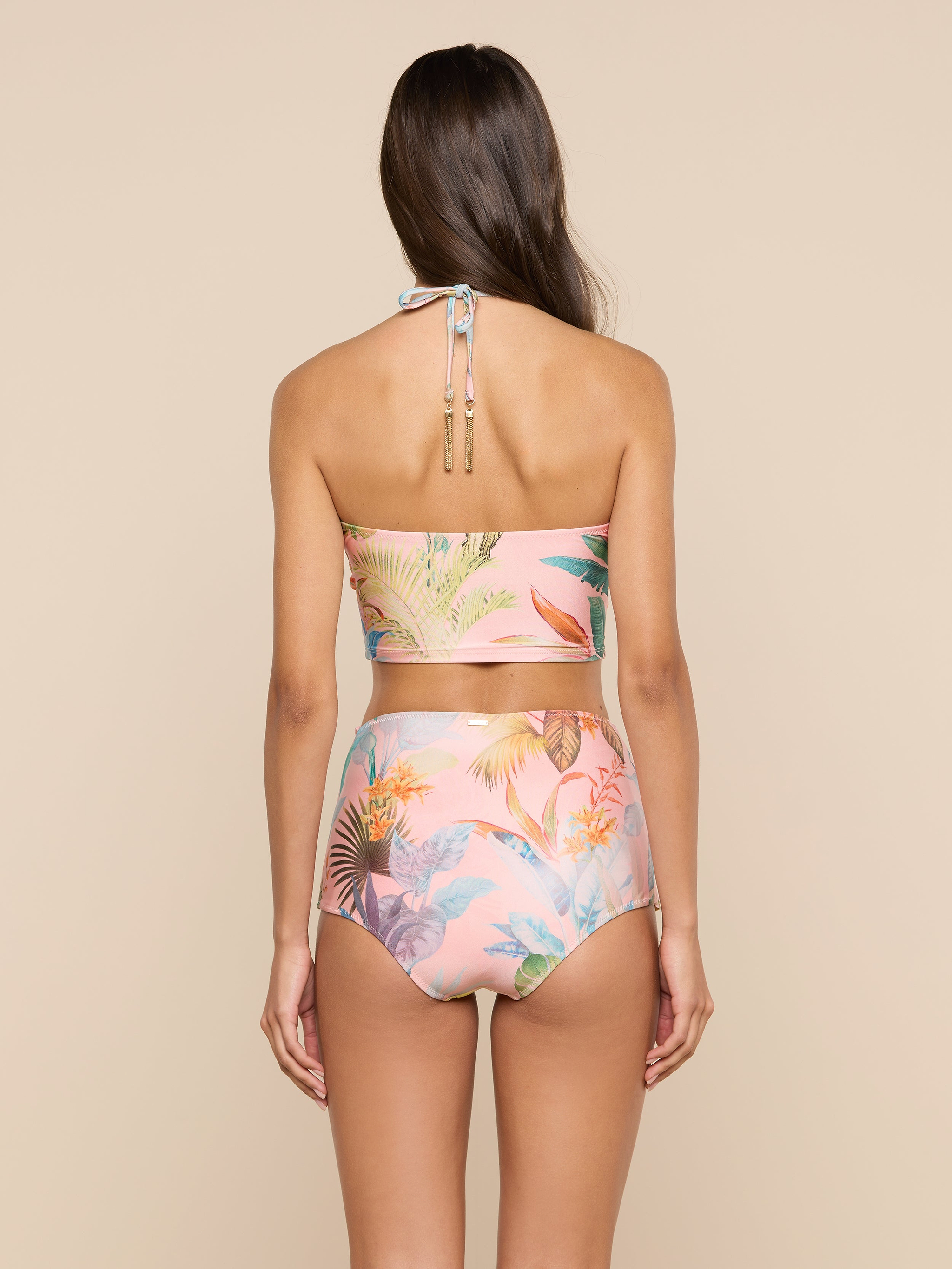 A woman with long brown hair is seen from the back, wearing the Siwan Midkini—a pastel floral two-piece swimsuit with a longline bikini top and high-waisted bottoms—standing against a plain light background.