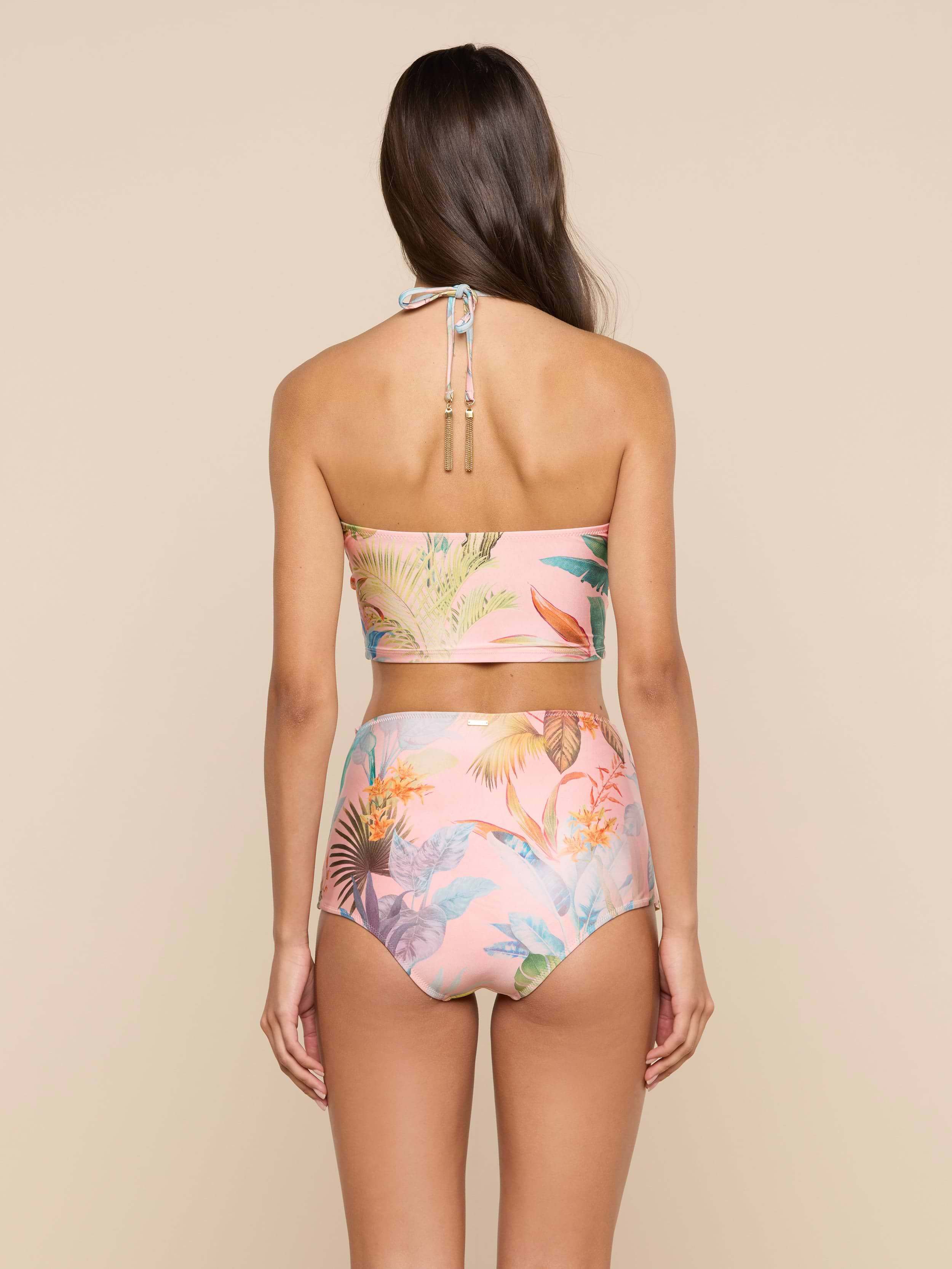 A woman with long brown hair is seen from the back, wearing the Siwan Midkini—a pastel floral two-piece swimsuit with a longline bikini top and high-waisted bottoms—standing against a plain light background.