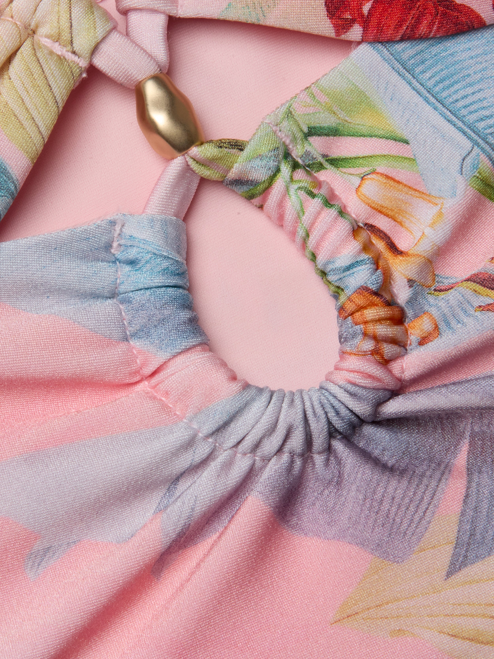 Close-up of the Siwan Midkini, showcasing colorful, patterned fabric with gathered elastic details and a gold bead, displayed on a light pink background. The midkini features soft pastel shades with a floral or abstract design.