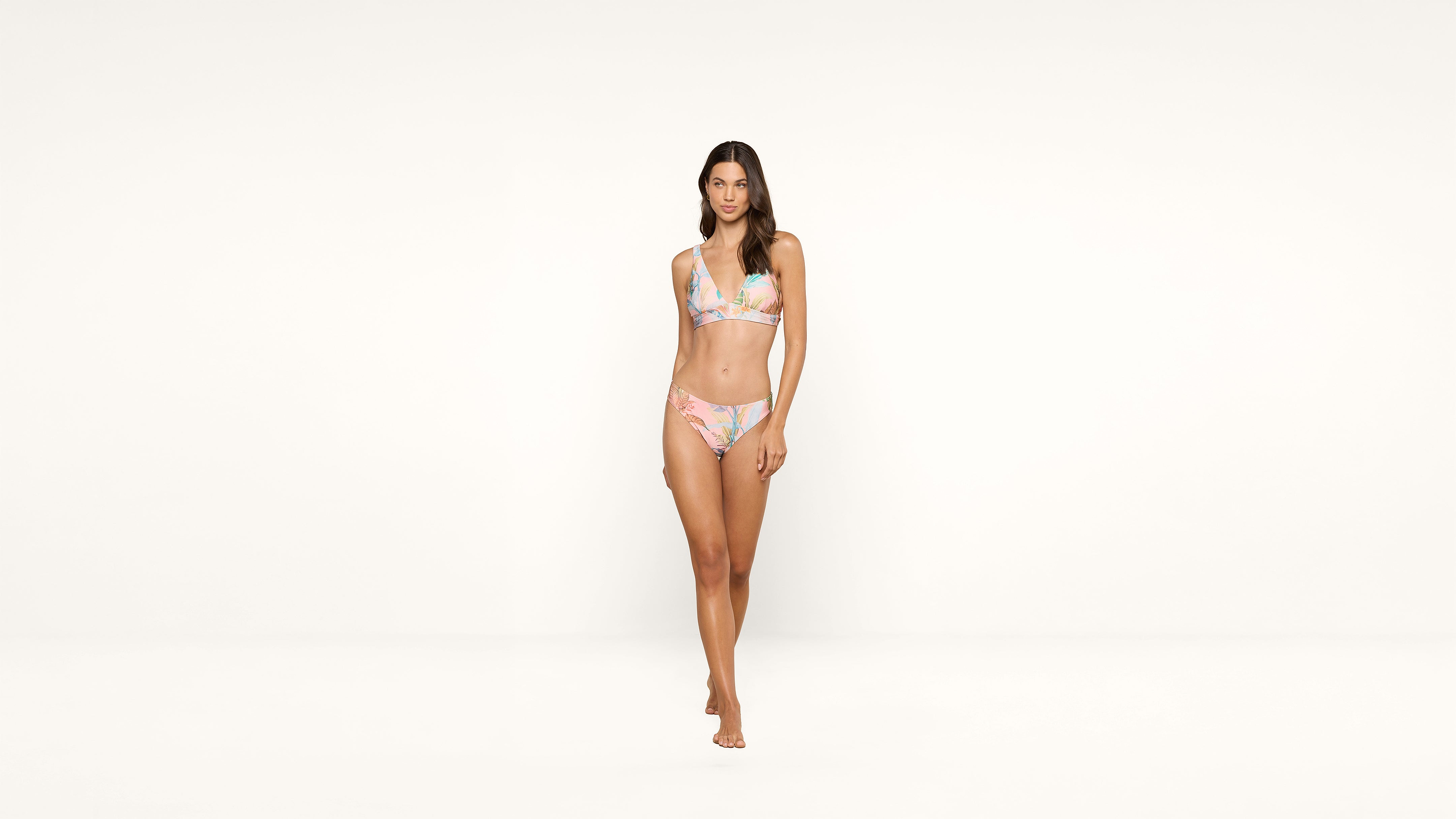 A woman wearing a pastel floral bikini stands barefoot against a plain white background, looking forward with a neutral expression.