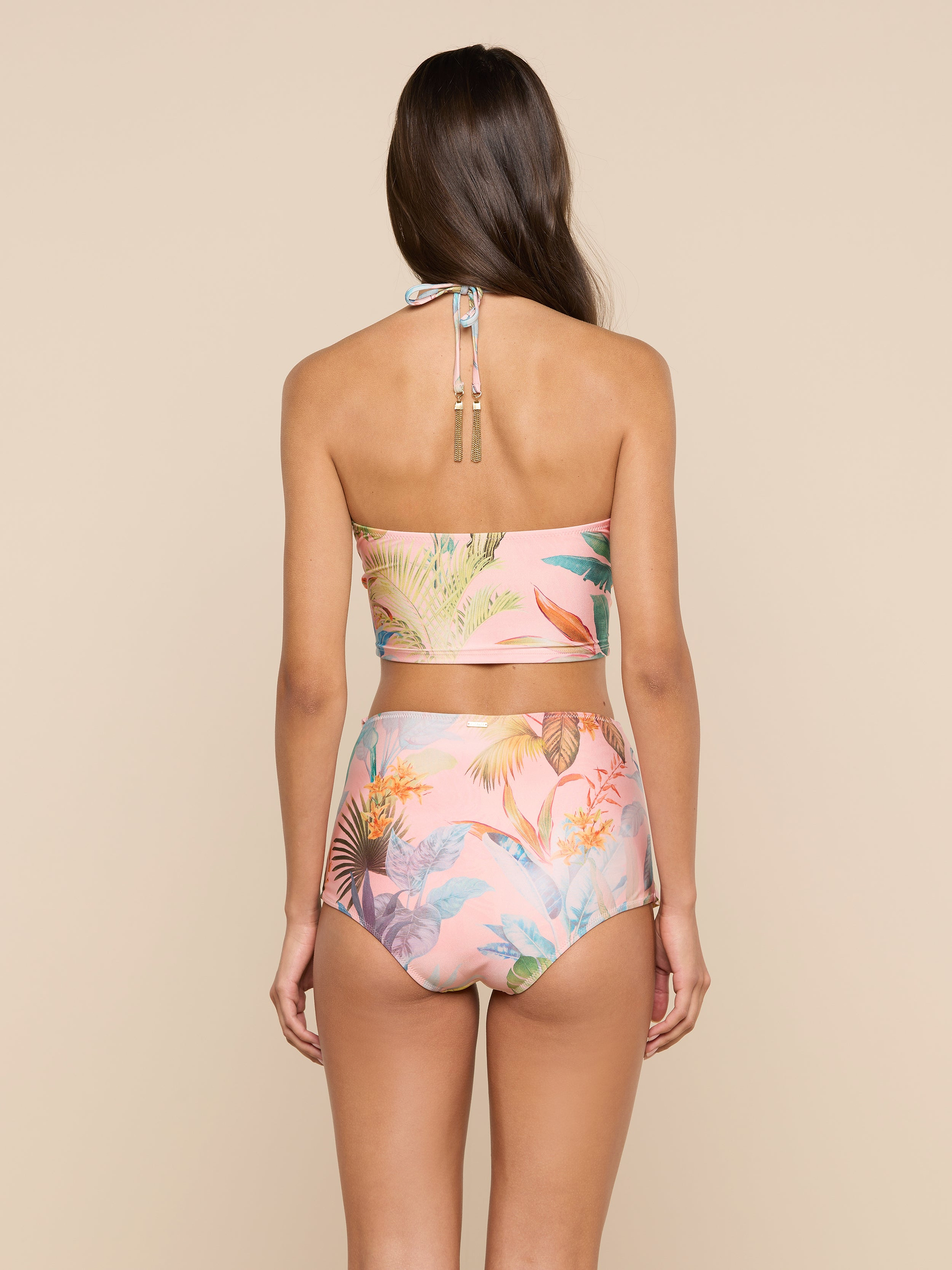 A woman with long brown hair, seen from the back, wears a pink, floral-patterned halter bikini top paired with the Linda Swim Skort. She stands against a plain, light background.