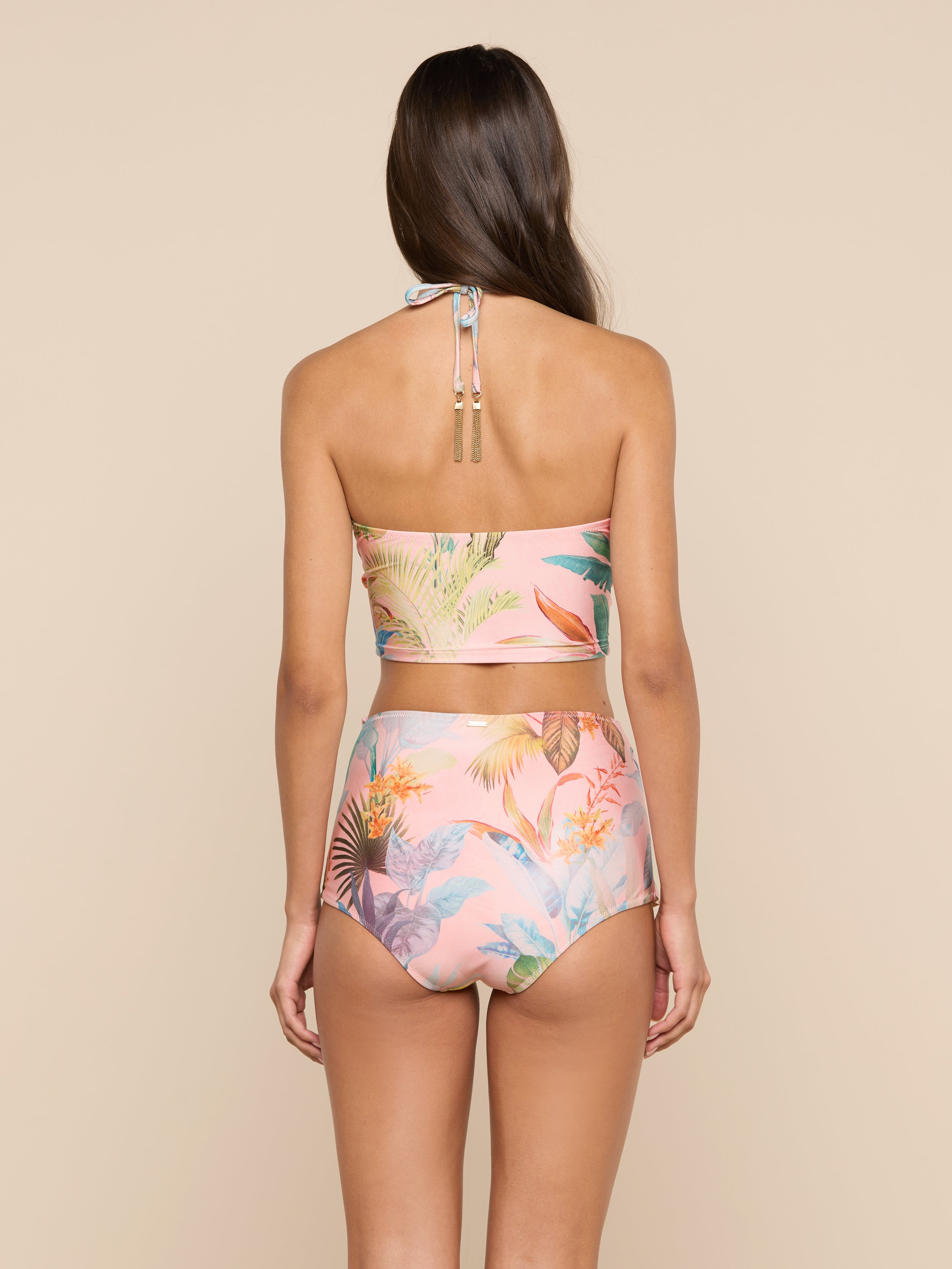 A woman with long brown hair, seen from the back, wears a pink, floral-patterned halter bikini top paired with the Linda Swim Skort. She stands against a plain, light background.