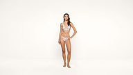 A woman stands barefoot against a plain white background, wearing a pastel-colored bikini with a floral pattern. She has long hair and a relaxed pose, with one hand resting on her hip.