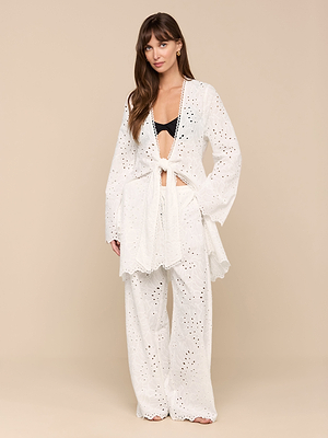 A woman stands against a beige background wearing the Navaeh Cover-Up Kimono with wide sleeves and scalloped edges over a black bra, paired with matching pants. Her long brown hair is styled in loose waves.
