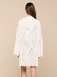 A woman with long brown hair, seen from behind, wears the Navaeh Cover-Up Kimono—a white, long-sleeve, knee-length cotton eyelet dress featuring a scalloped hem—against a plain light background.