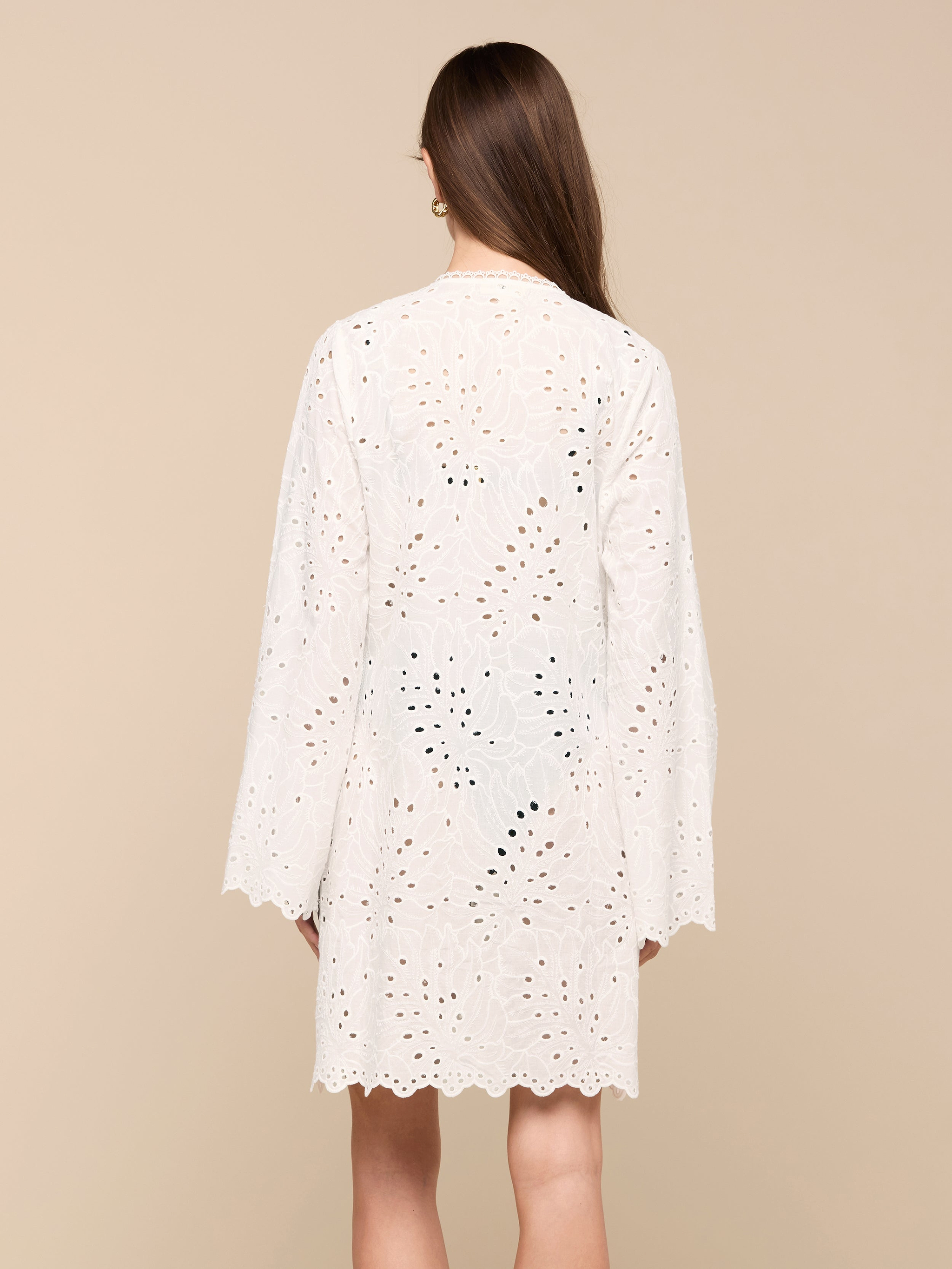 A woman with long brown hair, seen from behind, wears the Navaeh Cover-Up Kimono—a white, long-sleeve, knee-length cotton eyelet dress featuring a scalloped hem—against a plain light background.