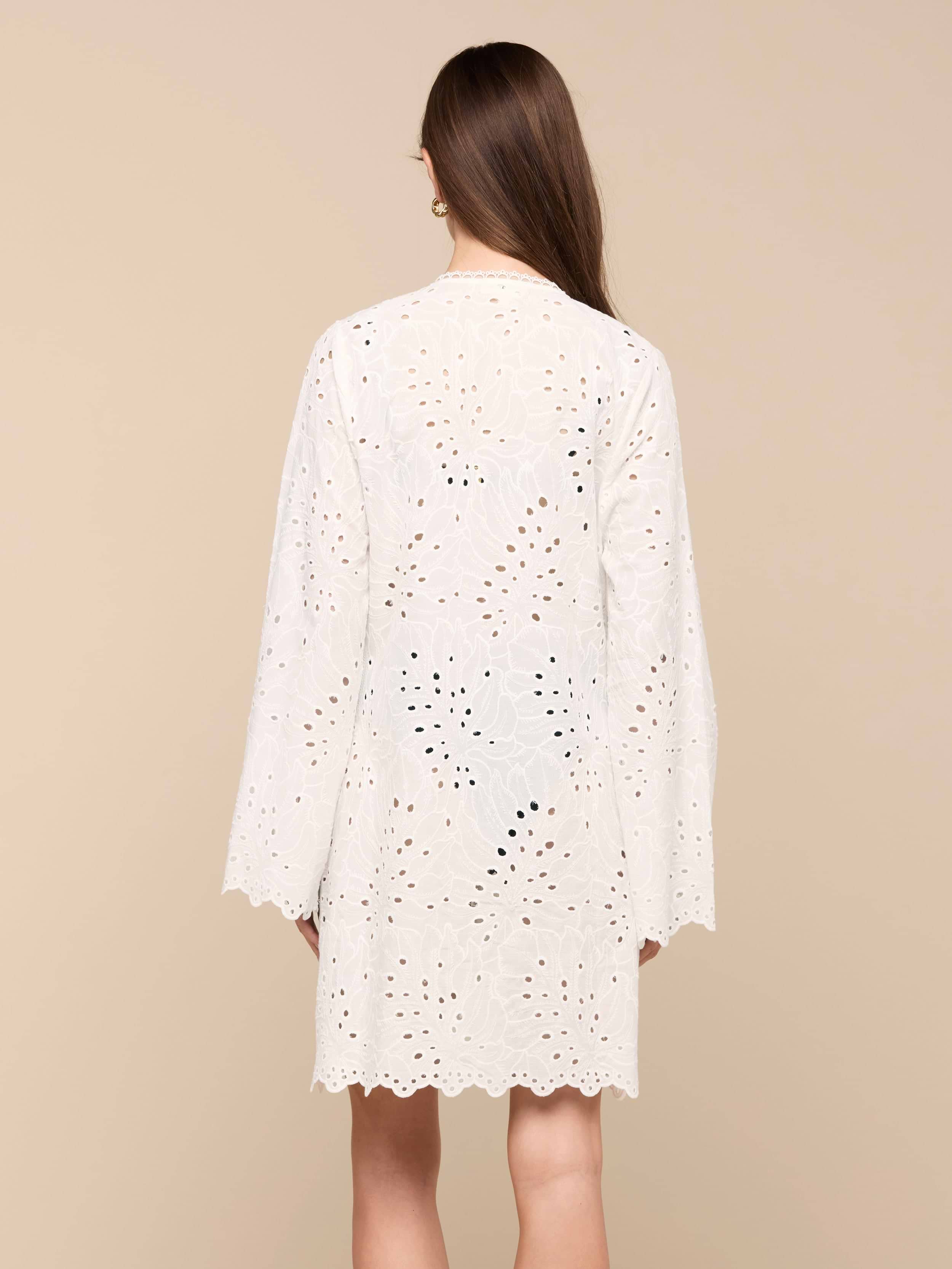 A woman with long brown hair, seen from behind, wears the Navaeh Cover-Up Kimono—a white, long-sleeve, knee-length cotton eyelet dress featuring a scalloped hem—against a plain light background.