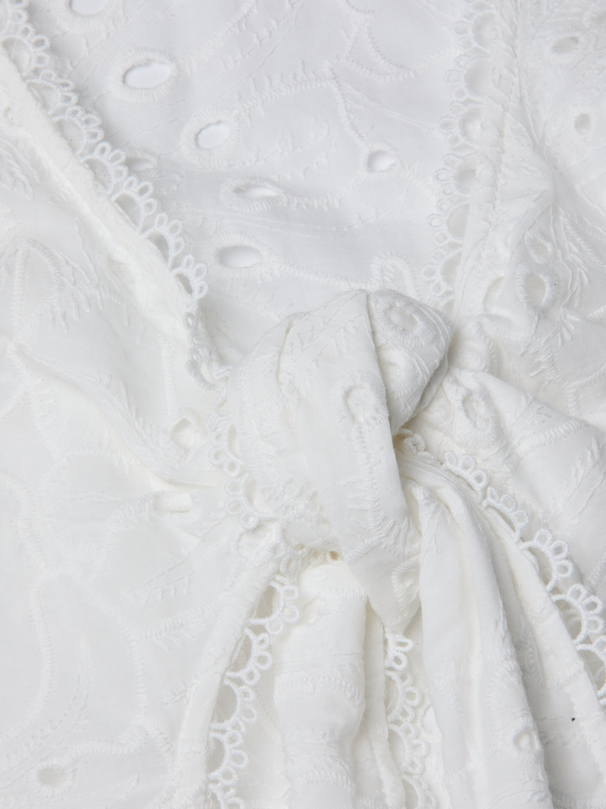 Close-up of the Navaeh Cover-Up Kimono, featuring white embroidered lace fabric tied in a knot, with delicate floral patterns and scalloped edges reminiscent of lightweight cotton eyelet.