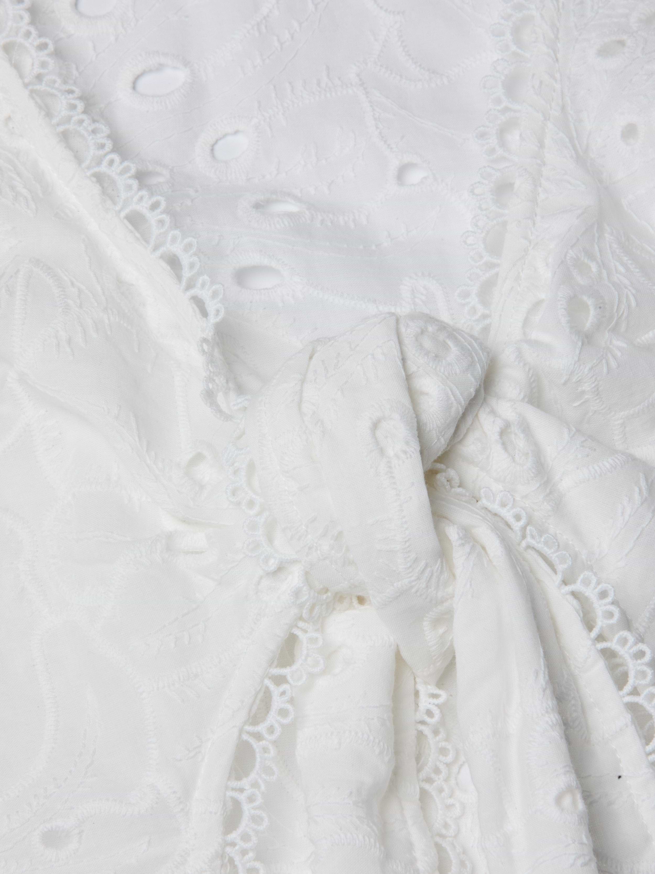 Close-up of the Navaeh Cover-Up Kimono, featuring white embroidered lace fabric tied in a knot, with delicate floral patterns and scalloped edges reminiscent of lightweight cotton eyelet.