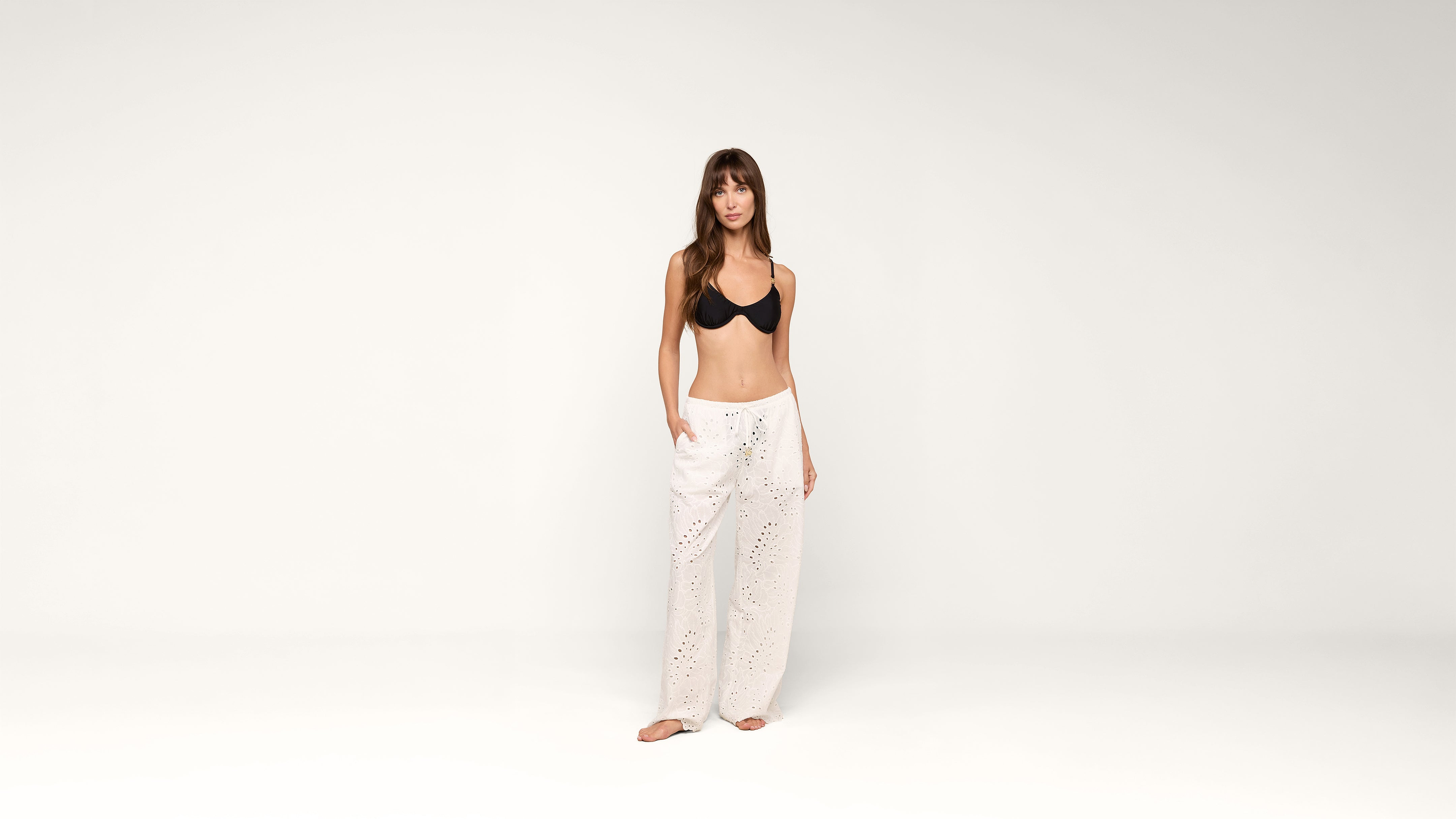 A woman stands barefoot against a plain white background, wearing a black bra and loose white eyelet pants with her hands in the pockets.