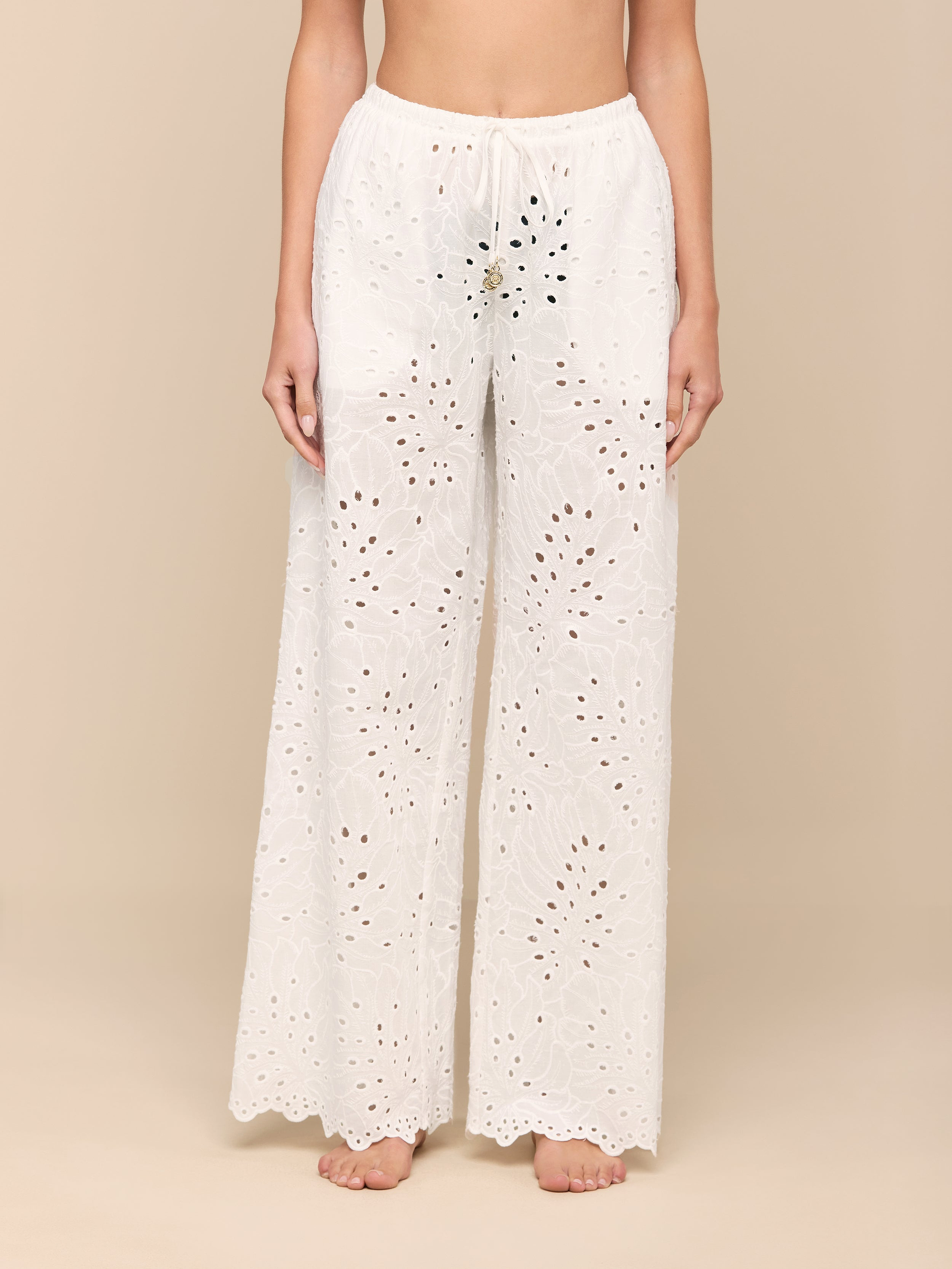 A person stands barefoot against a plain white background, wearing the Geraldine Eyelet Cover-Up Pant—a loose, white cotton style featuring an eyelet lace design, scalloped hem, and tie waist. The upper body and head are not visible.