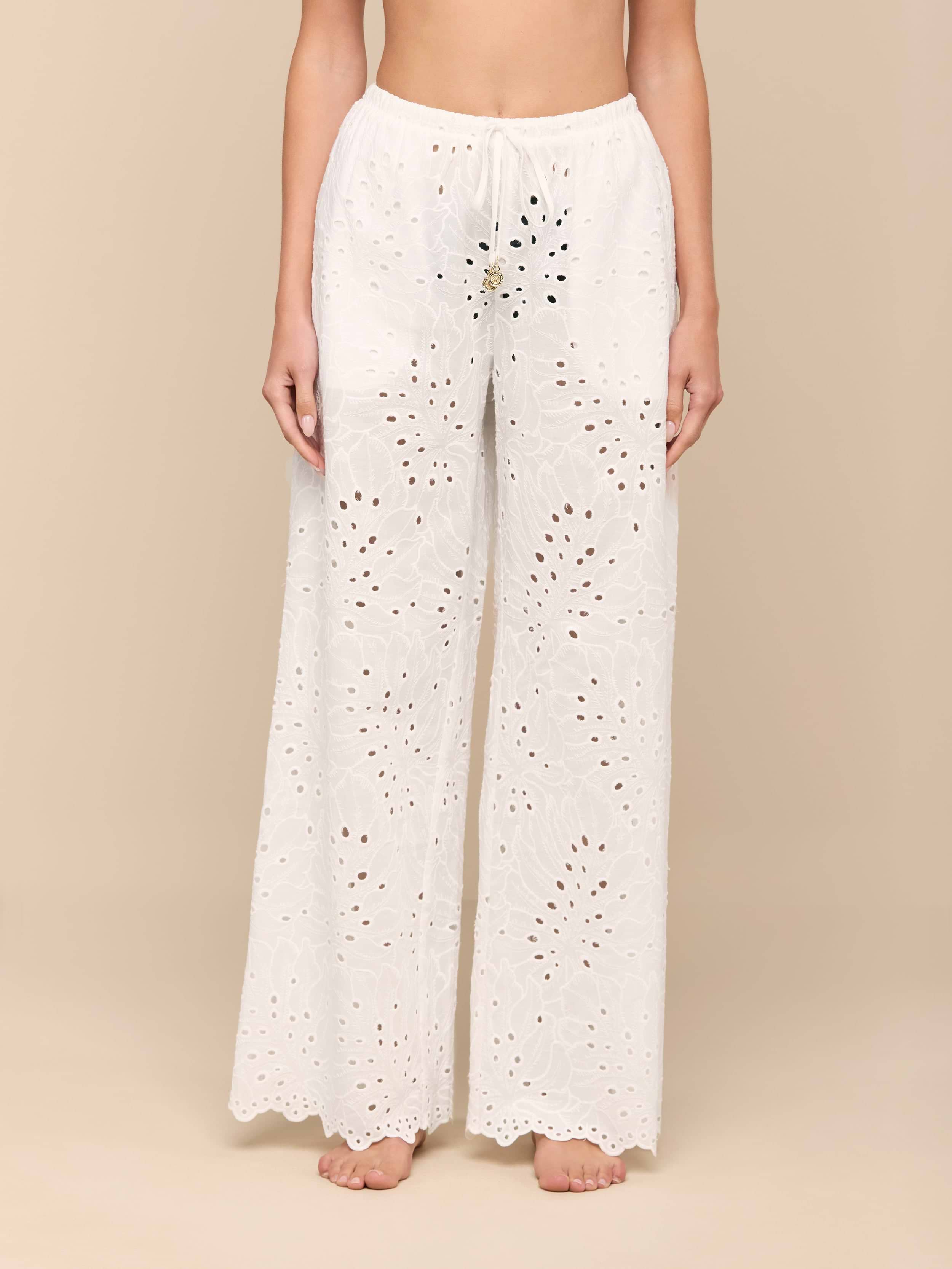 A person stands barefoot against a plain white background, wearing the Geraldine Eyelet Cover-Up Pant—a loose, white cotton style featuring an eyelet lace design, scalloped hem, and tie waist. The upper body and head are not visible.