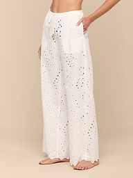 Barefoot, a person models the Geraldine Eyelet Cover-Up Pant—high-waisted, wide-leg cotton pants featuring a tie waist. The image focuses on the lower torso and legs set against a plain, light background.